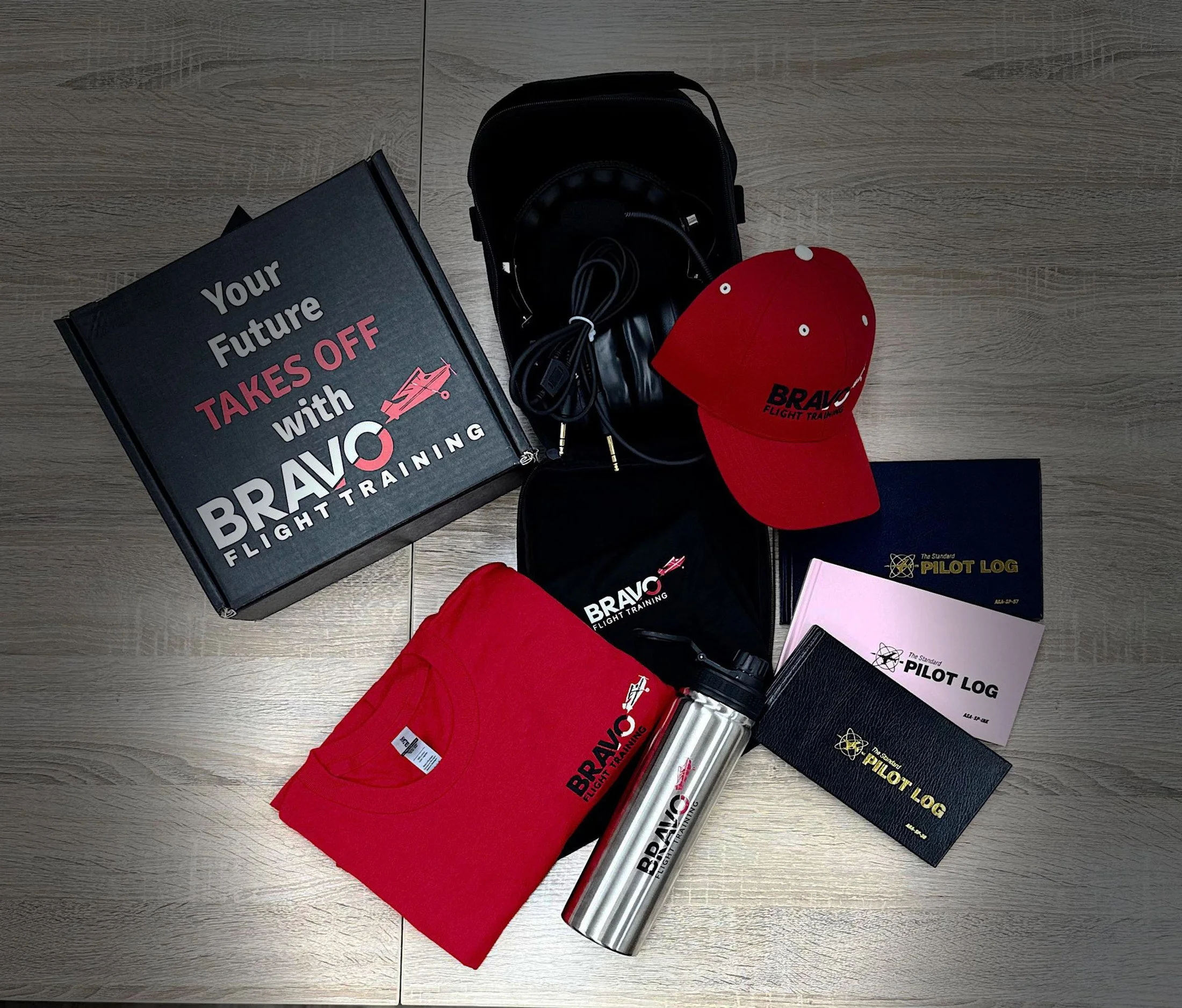 A collection of items related to aviation training, including a black book titled 'Your Future Takes Off with Bravo Flight Training,' a pair of black headphones with a microphone, a red baseball cap with the Bravo Flight Training logo, a stainless steel water bottle, pink and black notebooks labeled 'The Standard Pilot Log,' and a black booklet with a yellow logo, all arranged on a wooden surface.