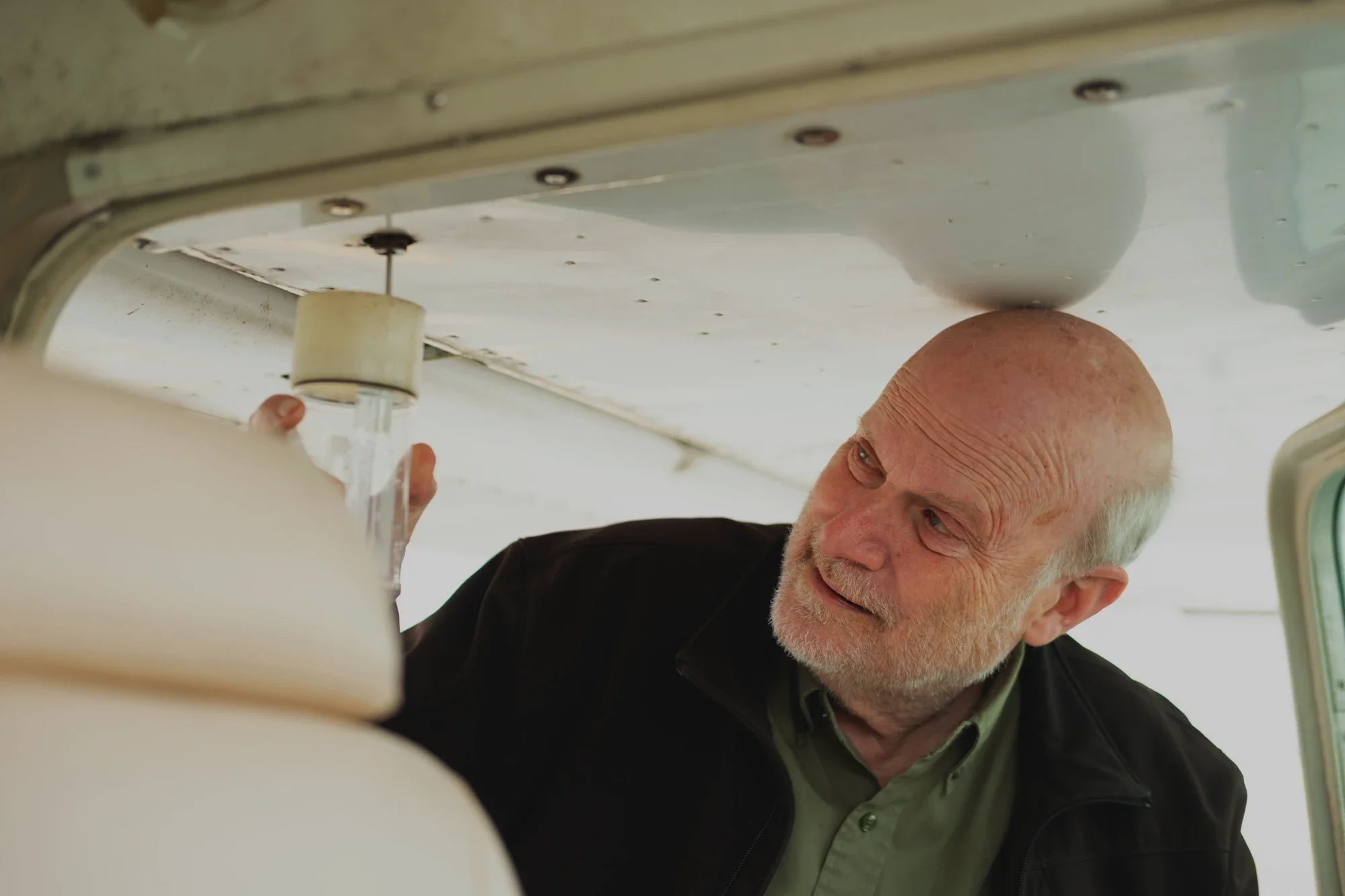An elderly man with a bald head and white facial hair is leaning over, looking at something out of frame inside an aircraft cabin with a white interior and overhead lighting.