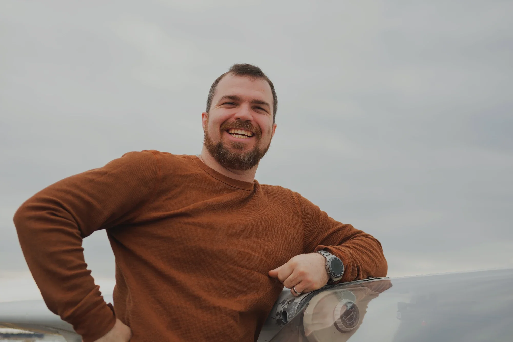 A smiling man with a beard and short hair leaning on an aircraft with a cloudy sky background.