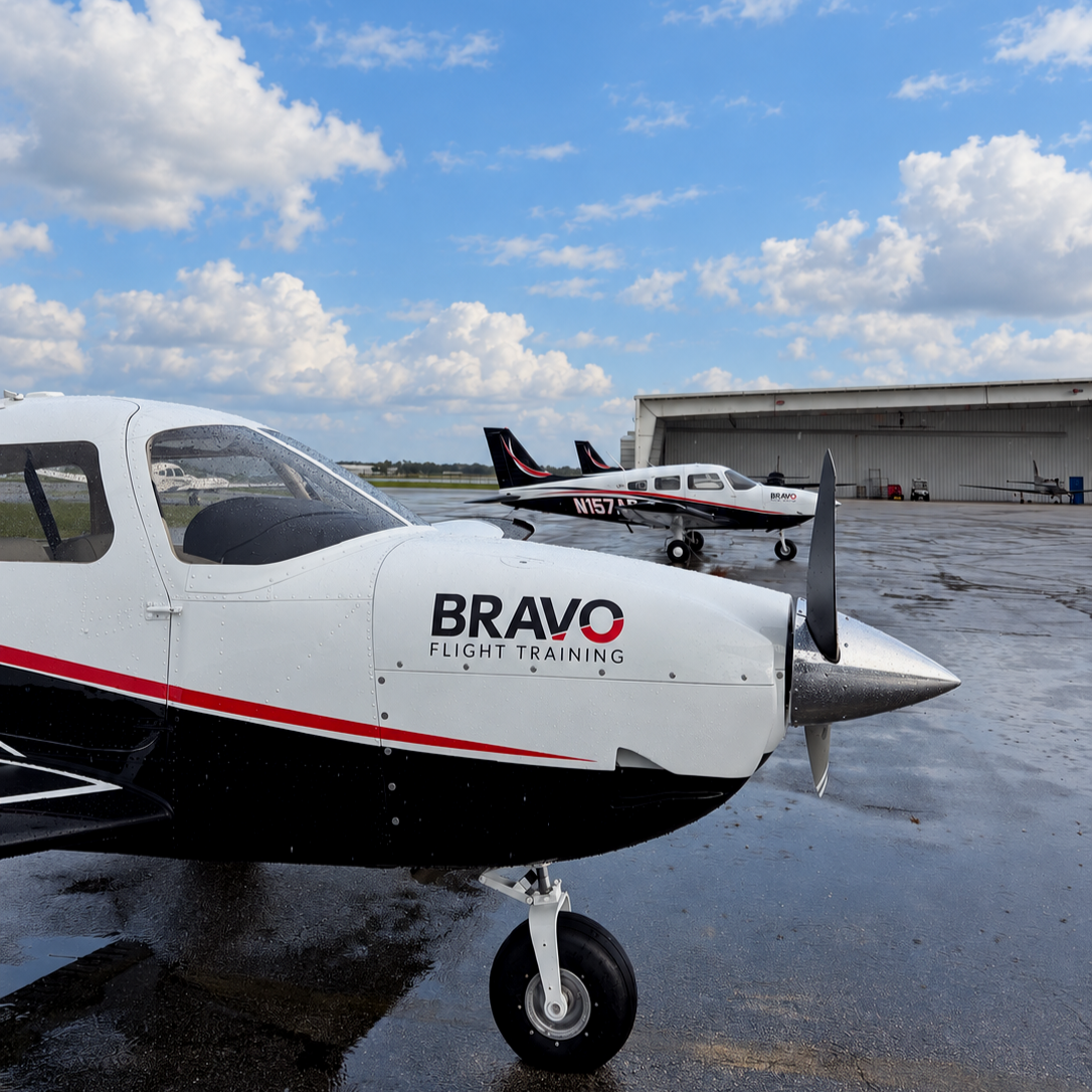 Small white airplane with Bravo Flight Training logo on the nose parked on a wet tarmac, with a hangar and another small airplane in the background under a partly cloudy sky.