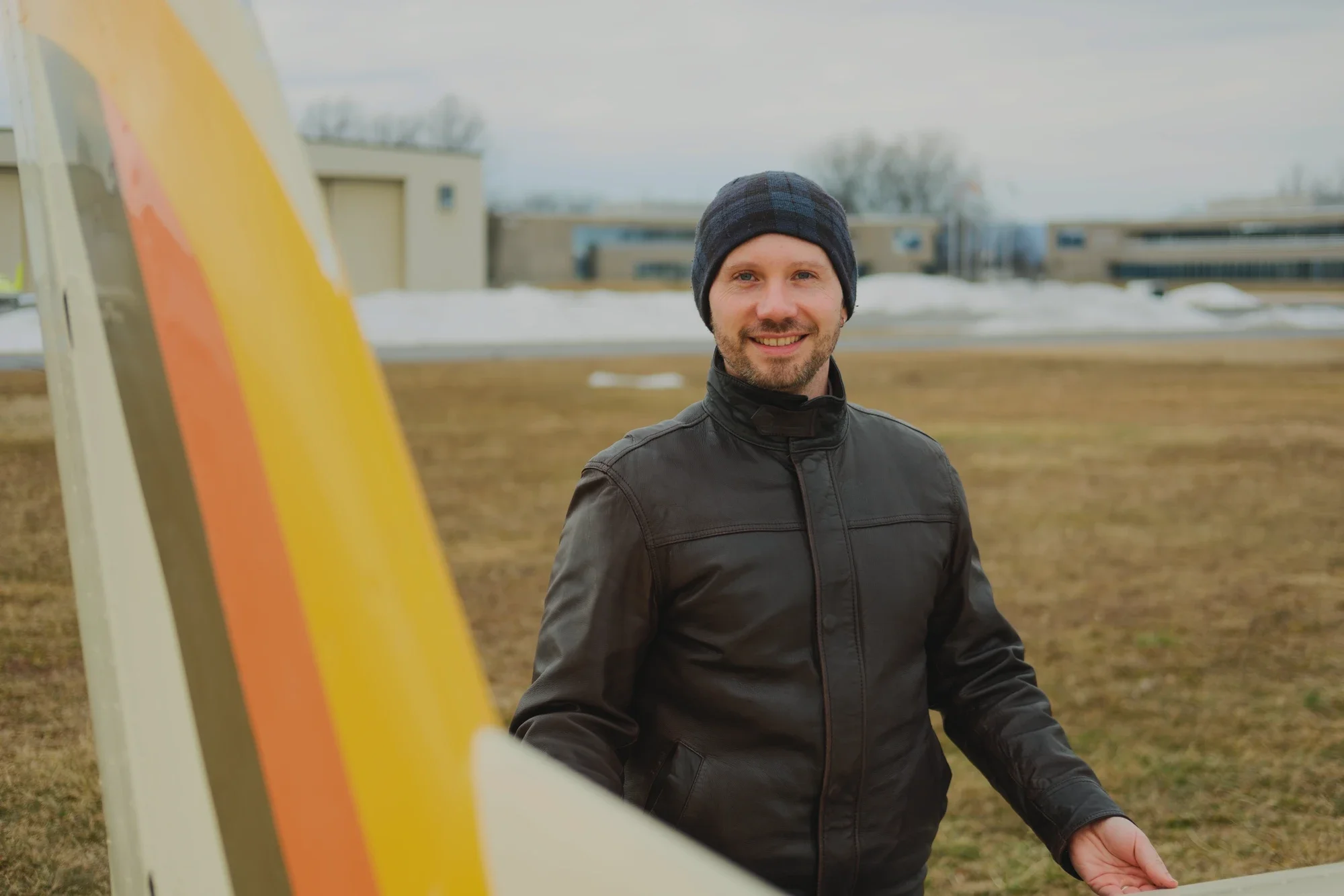 A smiling man wearing a black jacket and a navy blue beanie standing outdoors with snow and modern buildings in the background, partially obscured by a yellow and red structure in the foreground.