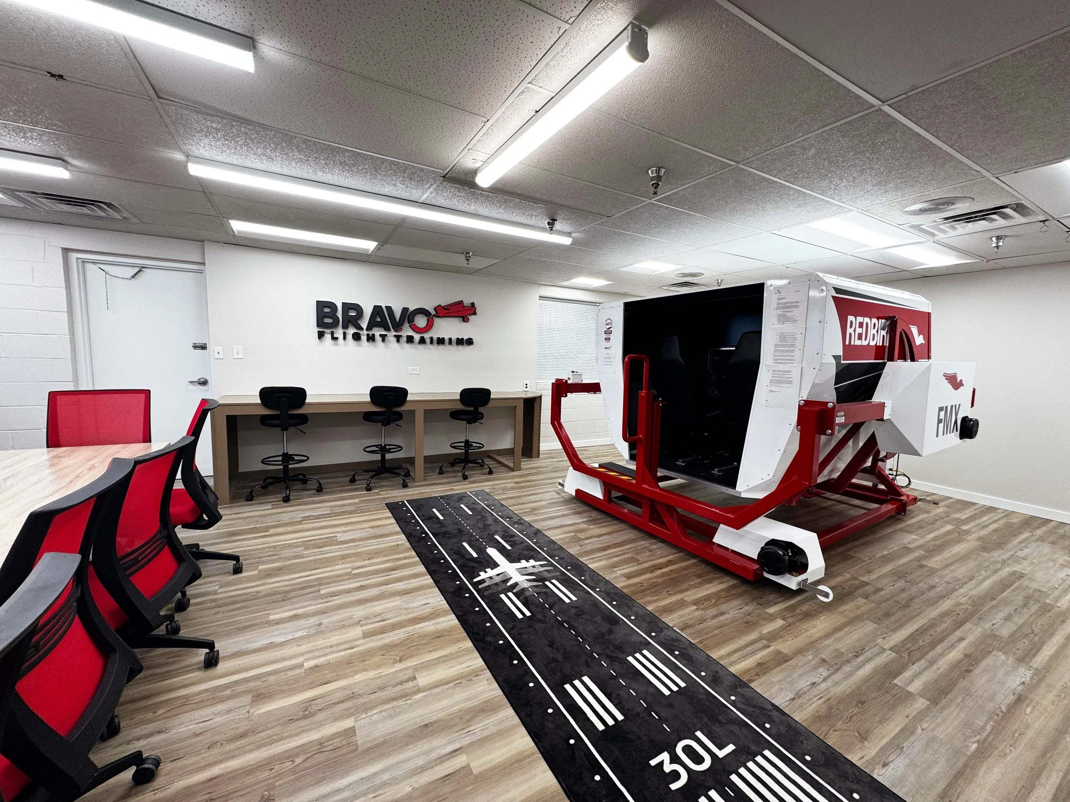 Simulator Room with Redbird FMX Full motion sim, Study table with red and black chairs, and a wall sign that says "BRAVO Flight Training."