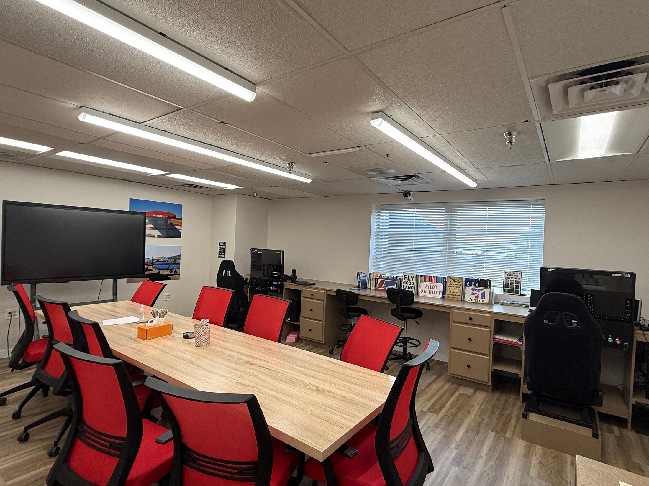 An office conference room with a large wooden table surrounded by red chairs, two TBD Flight simulators, a wall with books, posters, and a window with blinds, and a ceiling with fluorescent lights and ceiling tiles.