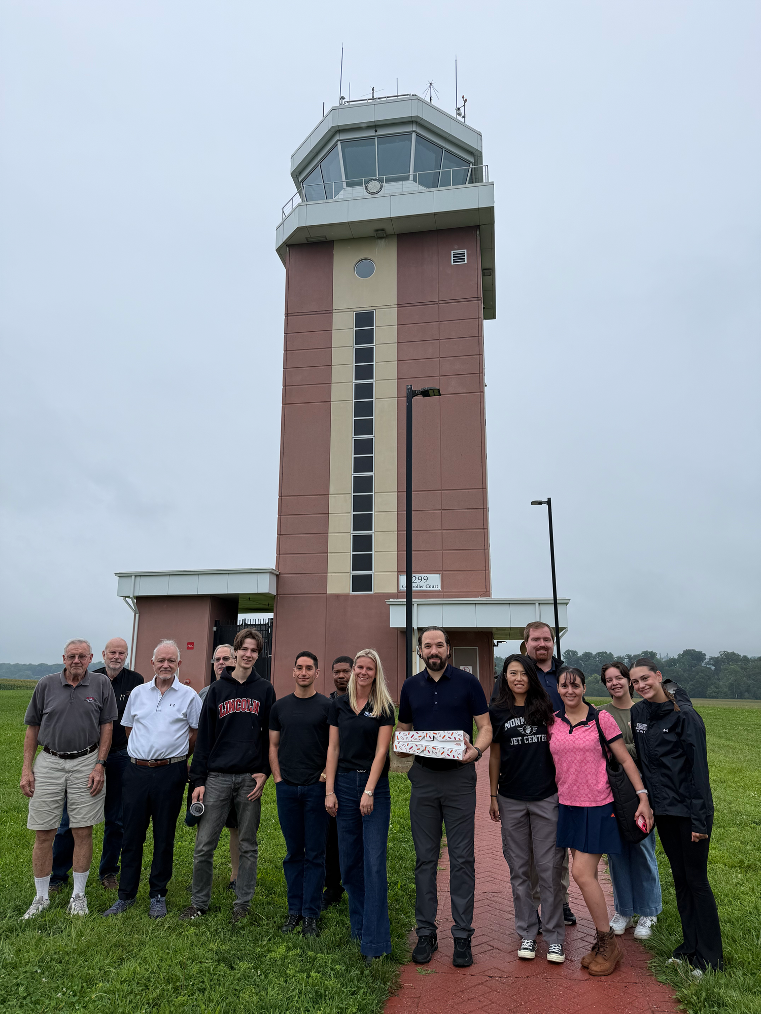 Group of people standing outdoors in front of an airport control tower, some smiling and holding a boxed gift.