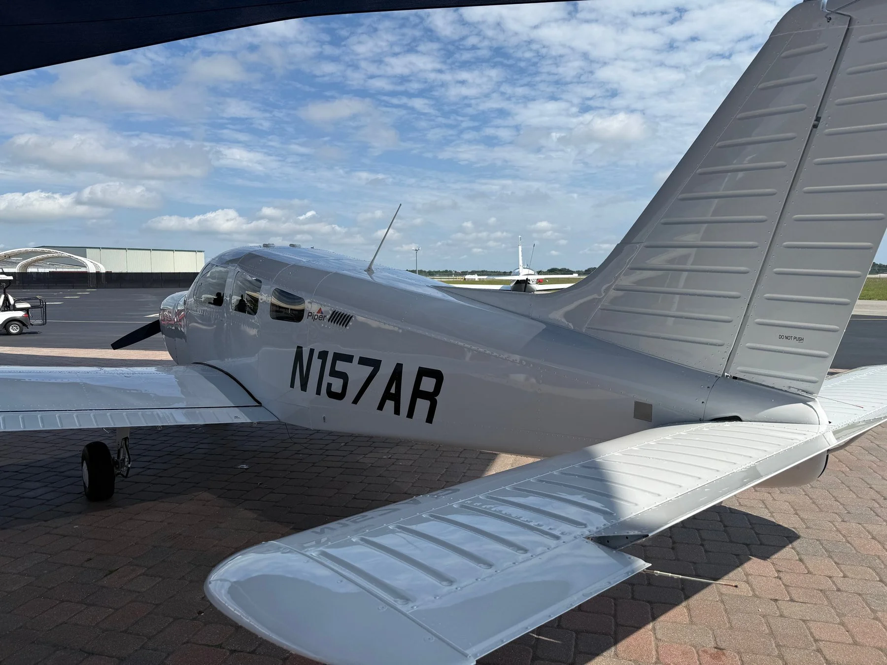 A small silver airplane parked on a tarmac at an airport, with a white hangar and a golf cart nearby, under a partly cloudy sky.