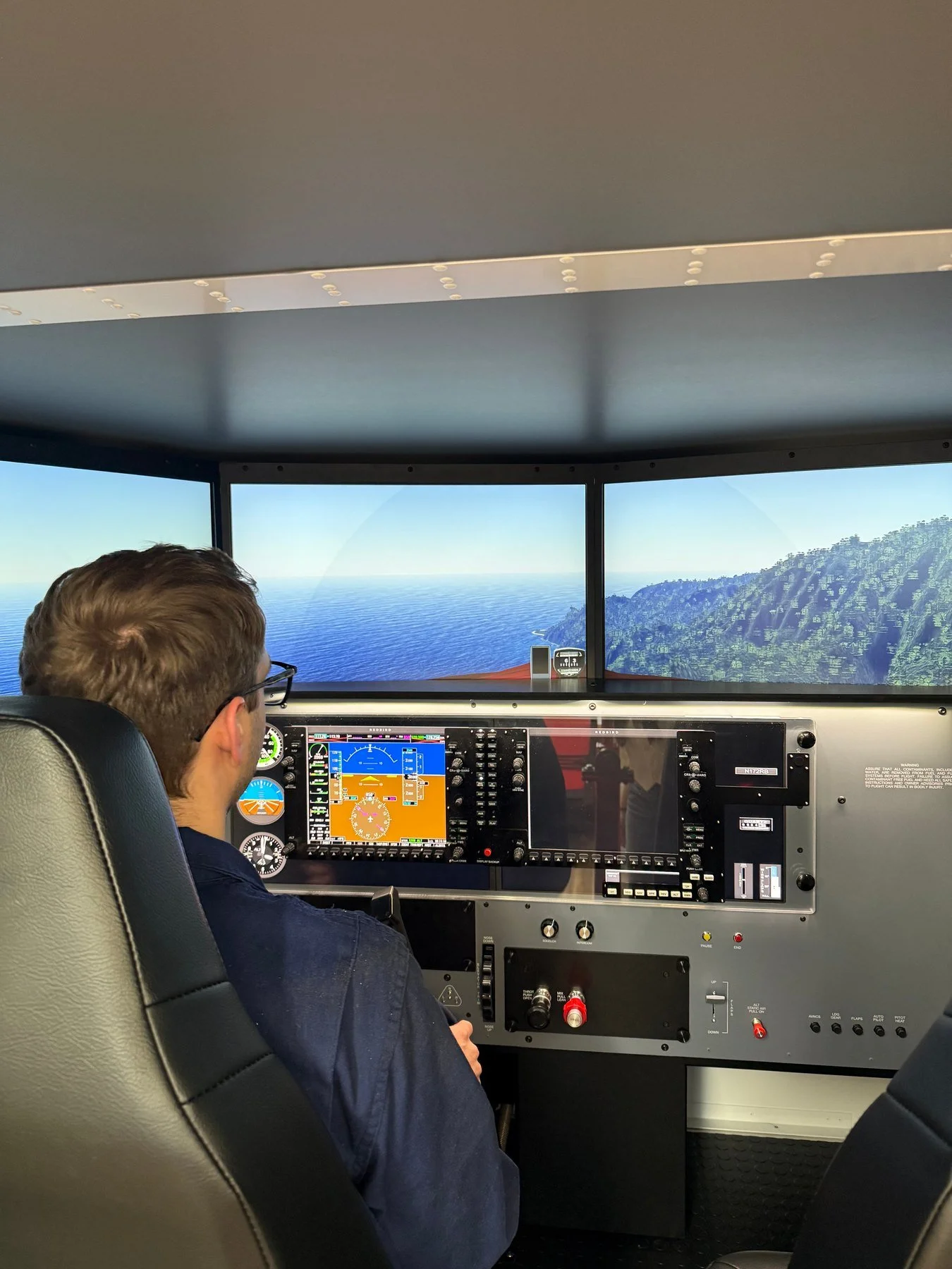 Pilot in cockpit of Redbird FMX Flight Simulator flying over ocean and coastline with mountains, view from pilot's seat showing aircraft instruments and screens, and window framing scenic landscape.