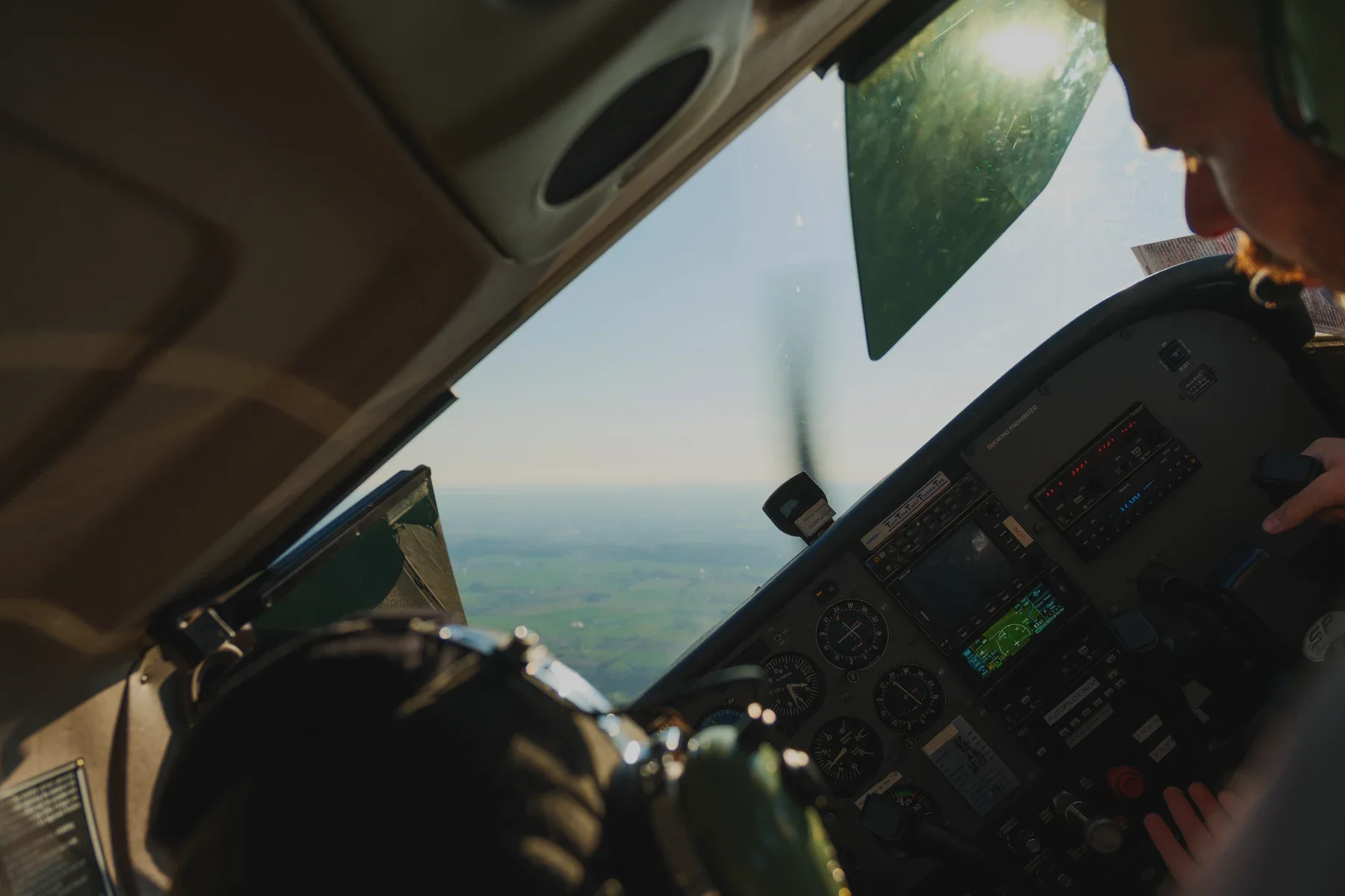 Inside the cockpit of an airplane showing the pilot and the instrument panel, view through the windshield of blue sky and green land below.