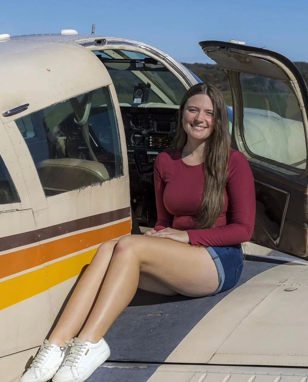 A woman with long brown hair sitting on the wing of a small beige airplane with orange and yellow stripes, smiling at the camera on a sunny day.
