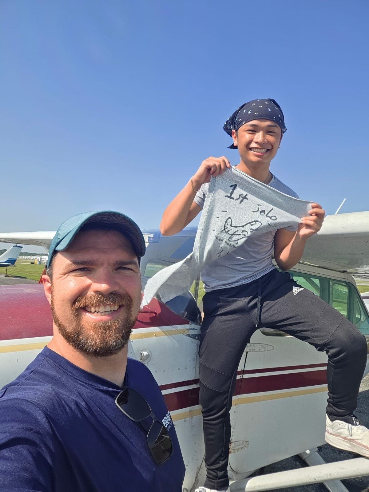 A smiling man with a beard and sunglasses hanging from his shirt taking a selfie in front of a red and white small airplane. A young man wearing a navy bandana and gray t-shirt holding a gray t-shirt with writing and a drawing, sitting on the airplane's wing, with a clear blue sky in the background.