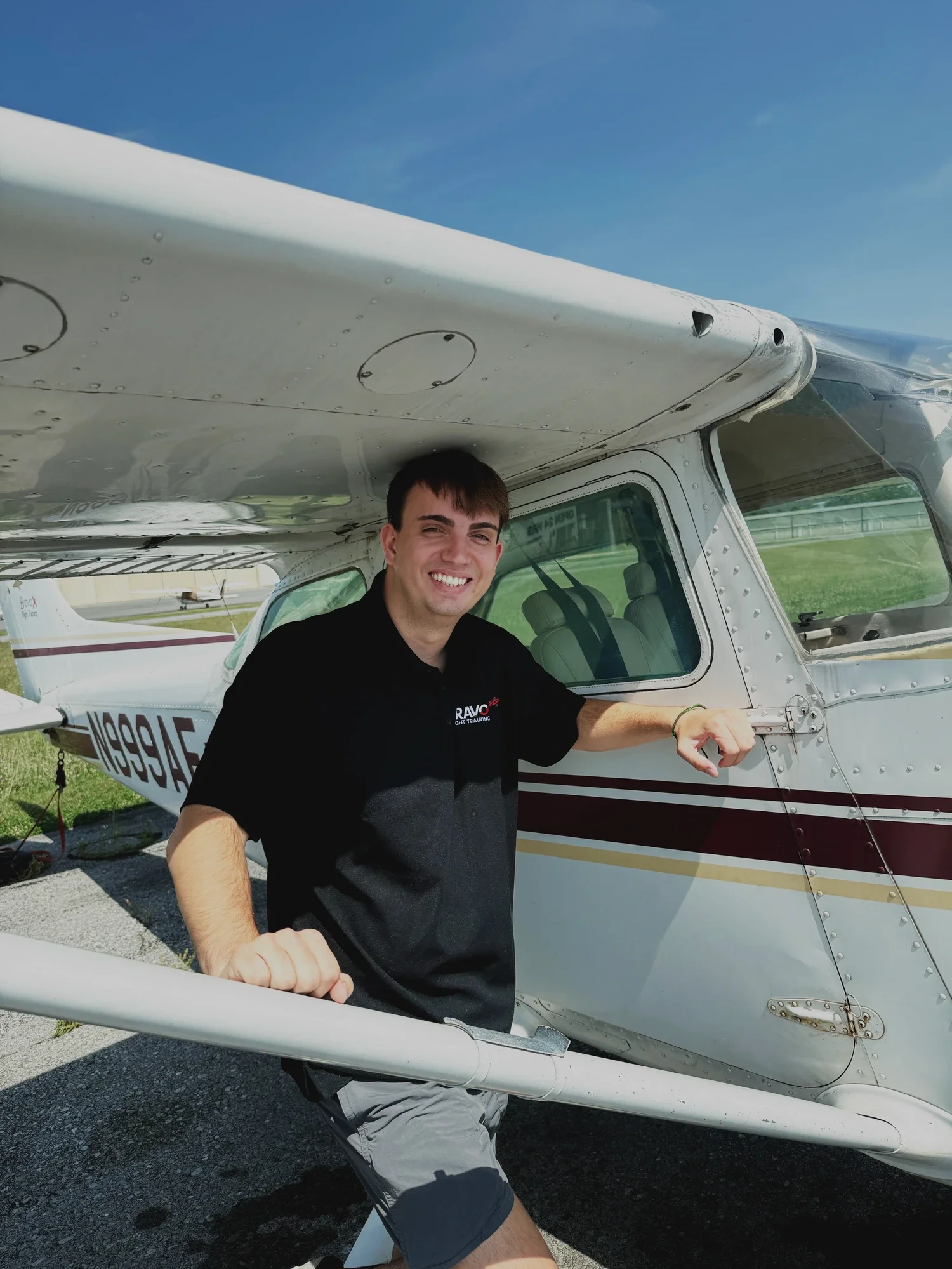 A smiling man in a black shirt and gray shorts standing next to a small white airplane with maroon and gold stripes, outdoors on a sunny day, with a blue sky and another small airplane in the background.