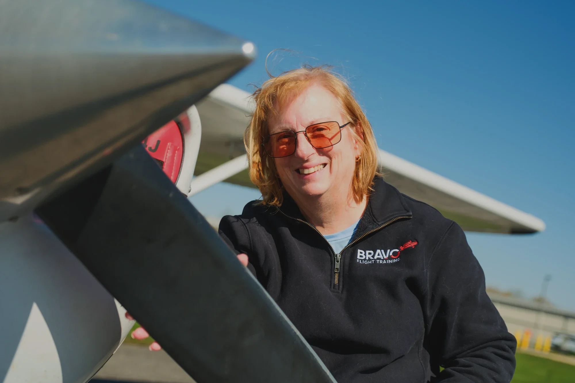 Woman with red hair and sunglasses smiling in front of an aircraft with the logo 'BRAVO Flight Training' on her black jacket, under a clear blue sky.
