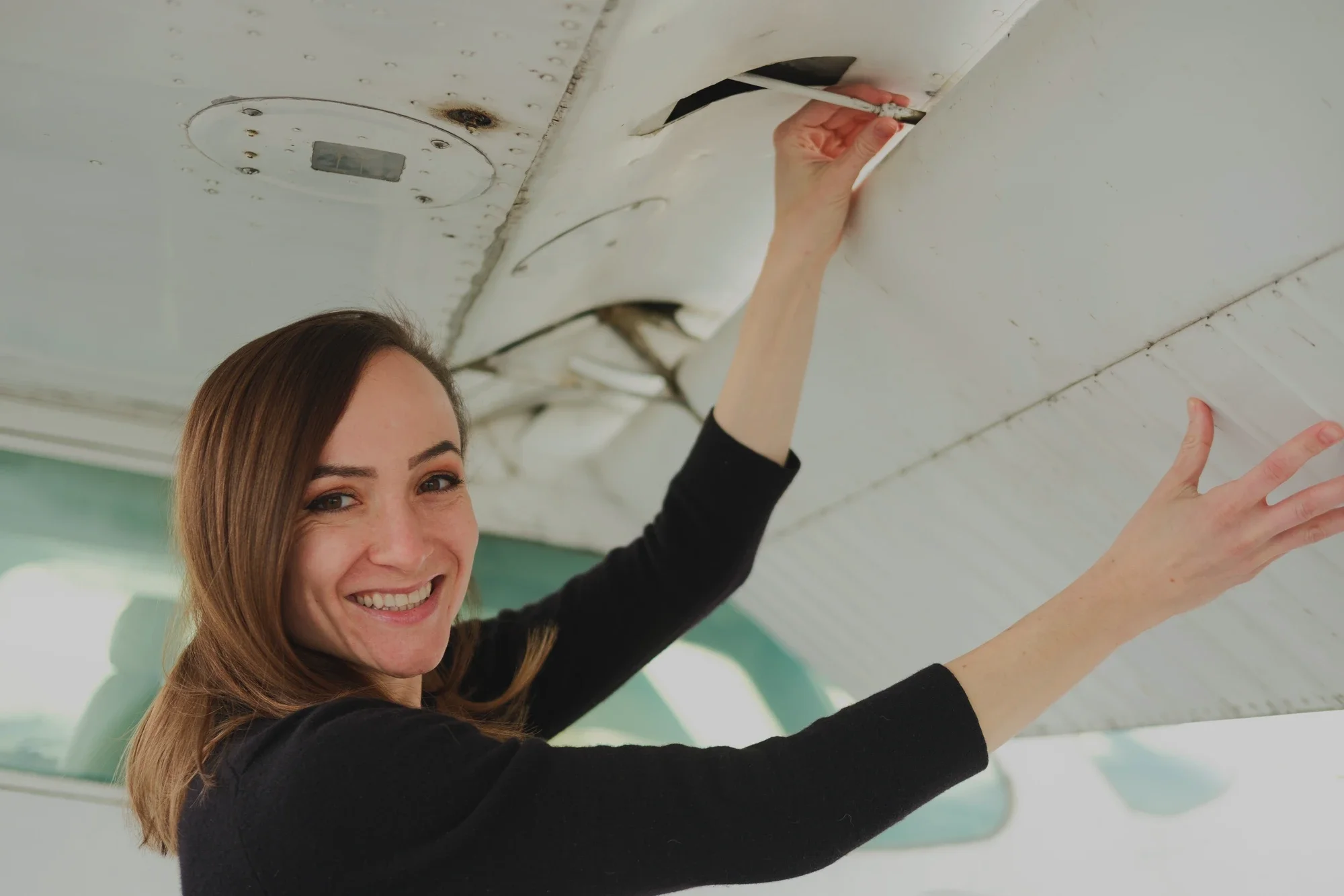 Woman with long brown hair smiling, fixing a ceiling light fixture in an indoor space.
