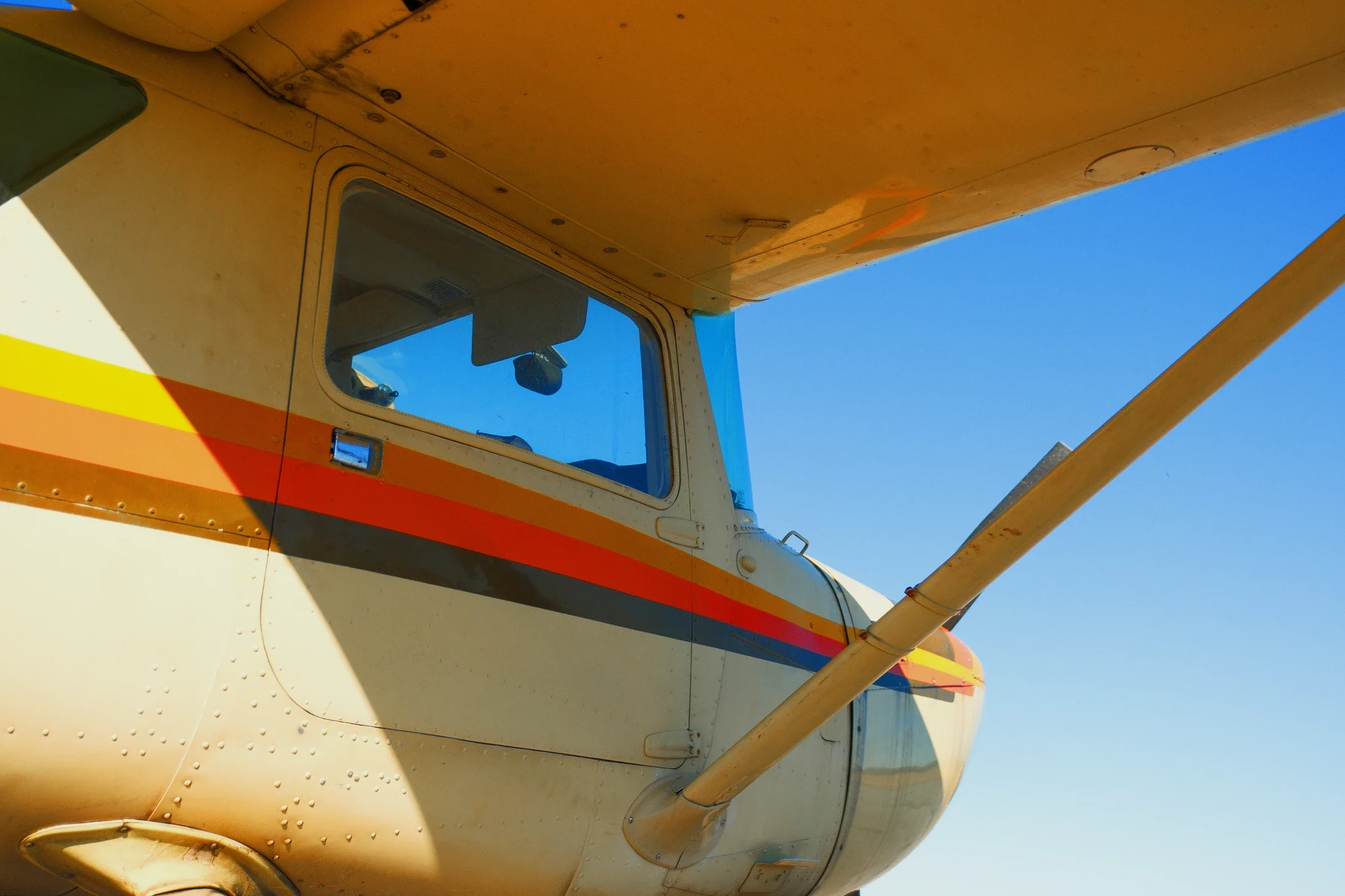 Close-up of the side of a small yellow aircraft with a red and orange stripe, showing the cockpit window, part of the wing, and the fuselage against a clear blue sky.