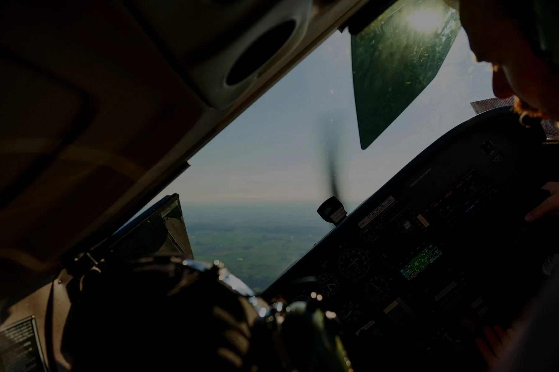 Inside the cockpit of an aircraft in flight, showing the pilot's hand on controls and a view of the landscape below through the windshield with a glare from the sun.