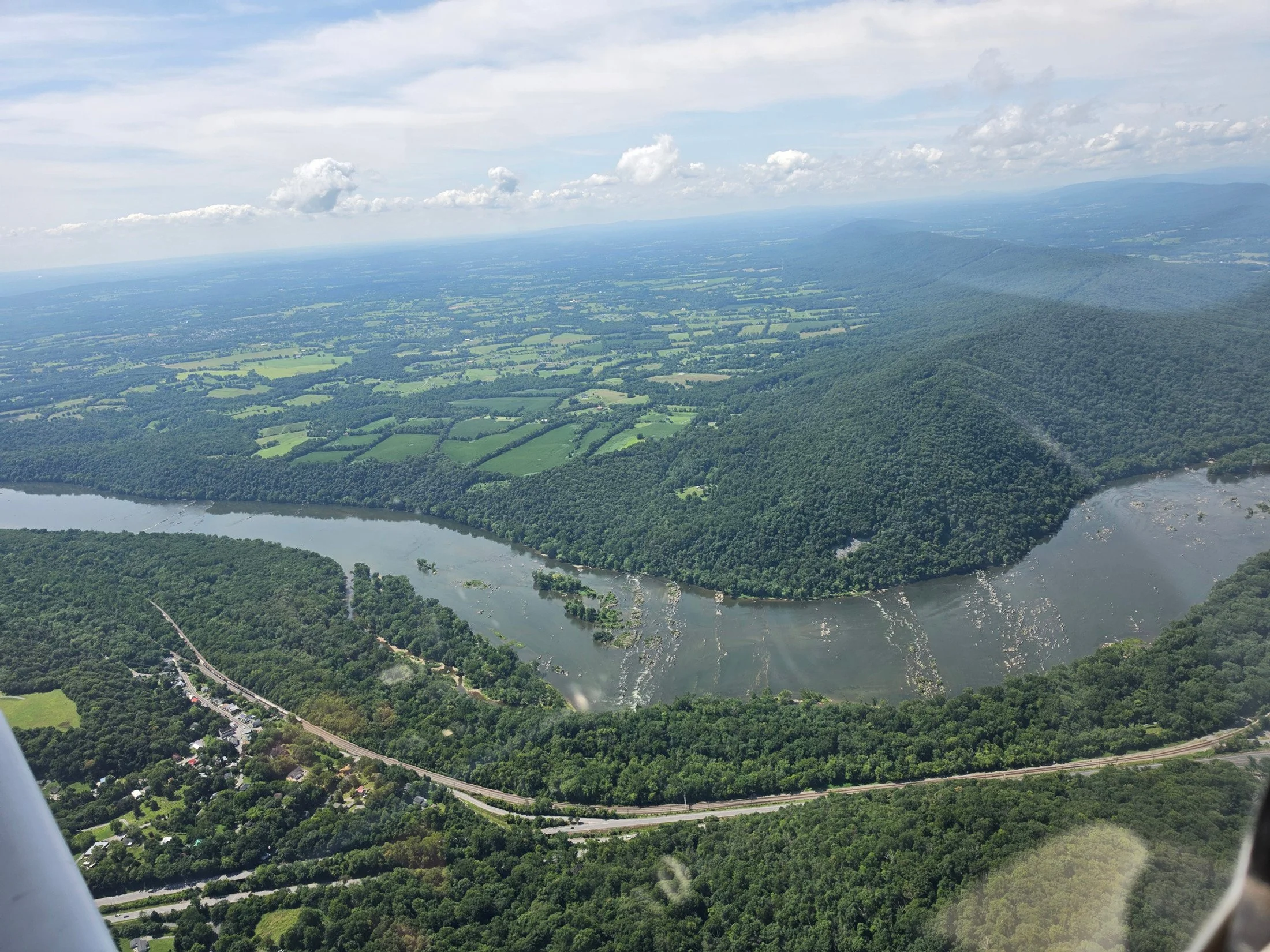 Aerial view of the potomac river winding through a green landscape with rolling hills, farmland, and a small town at the base of the hills, under a partly cloudy sky.