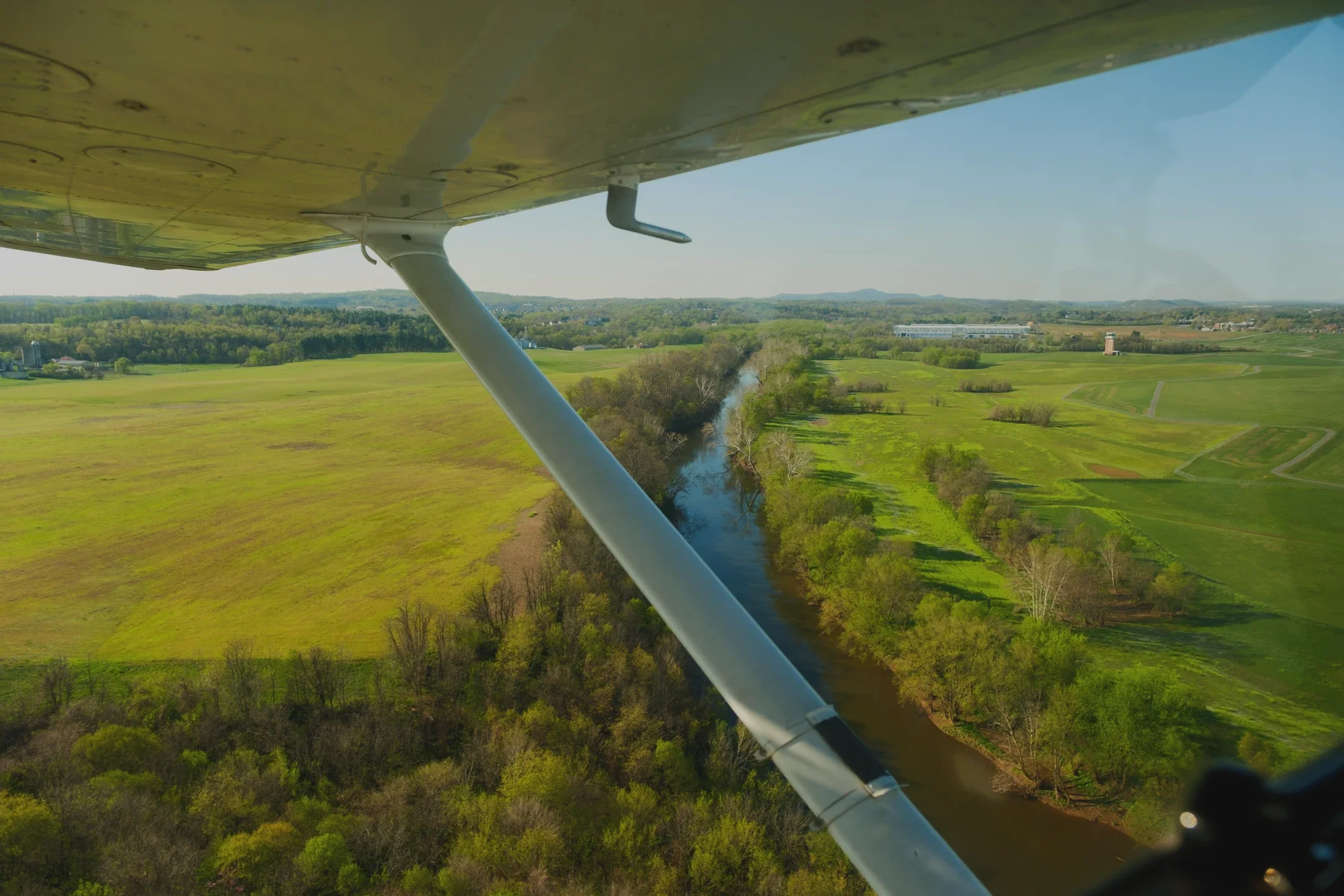 An aerial view from an airplane flying over a rural landscape with green fields, a river, and distant buildings under a clear sky.