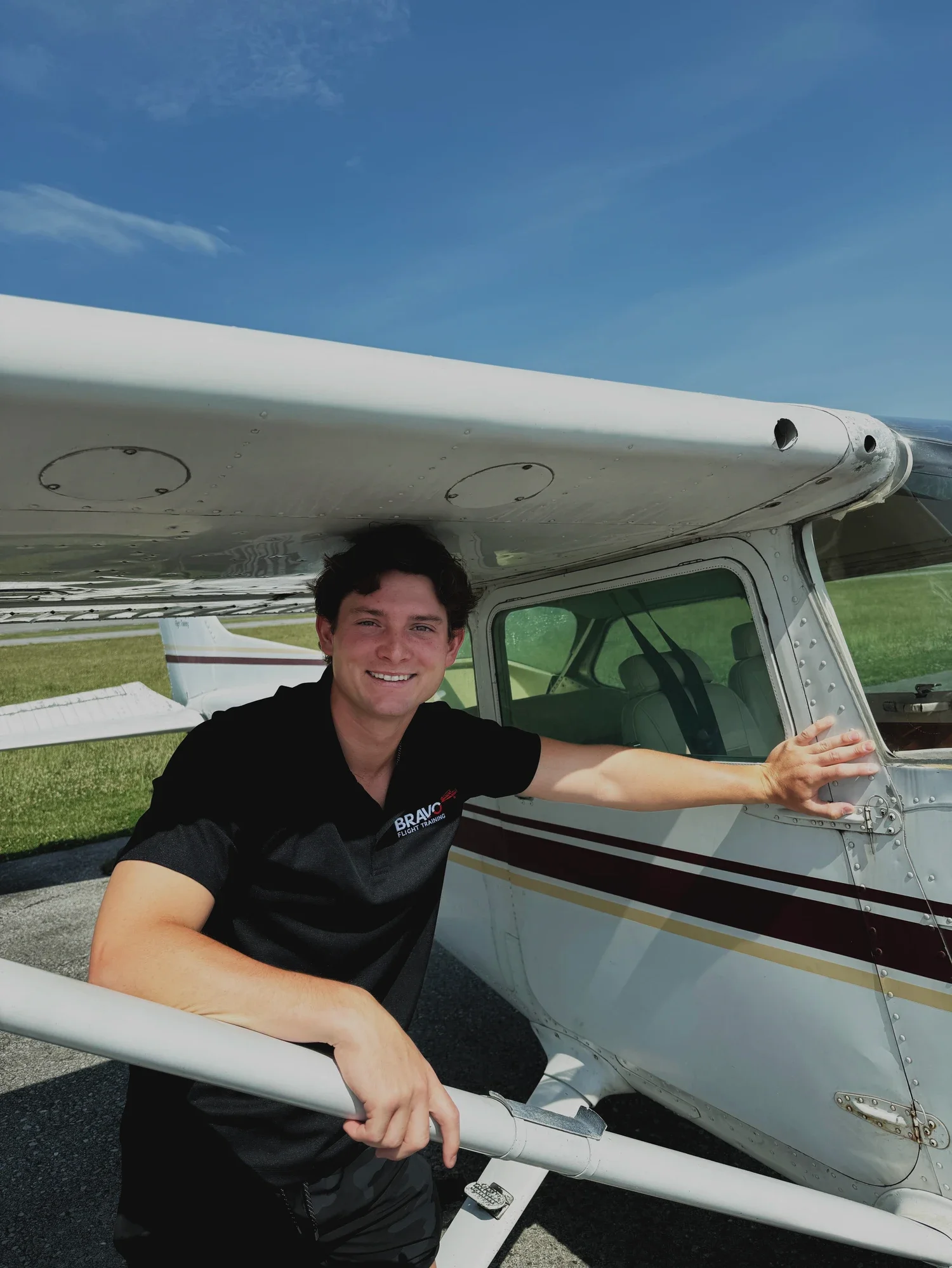 A young man smiling, wearing a black shirt, is standing next to a small white airplane. He is holding onto the airplane's wing, with a grassy field and a blue sky with some clouds in the background.