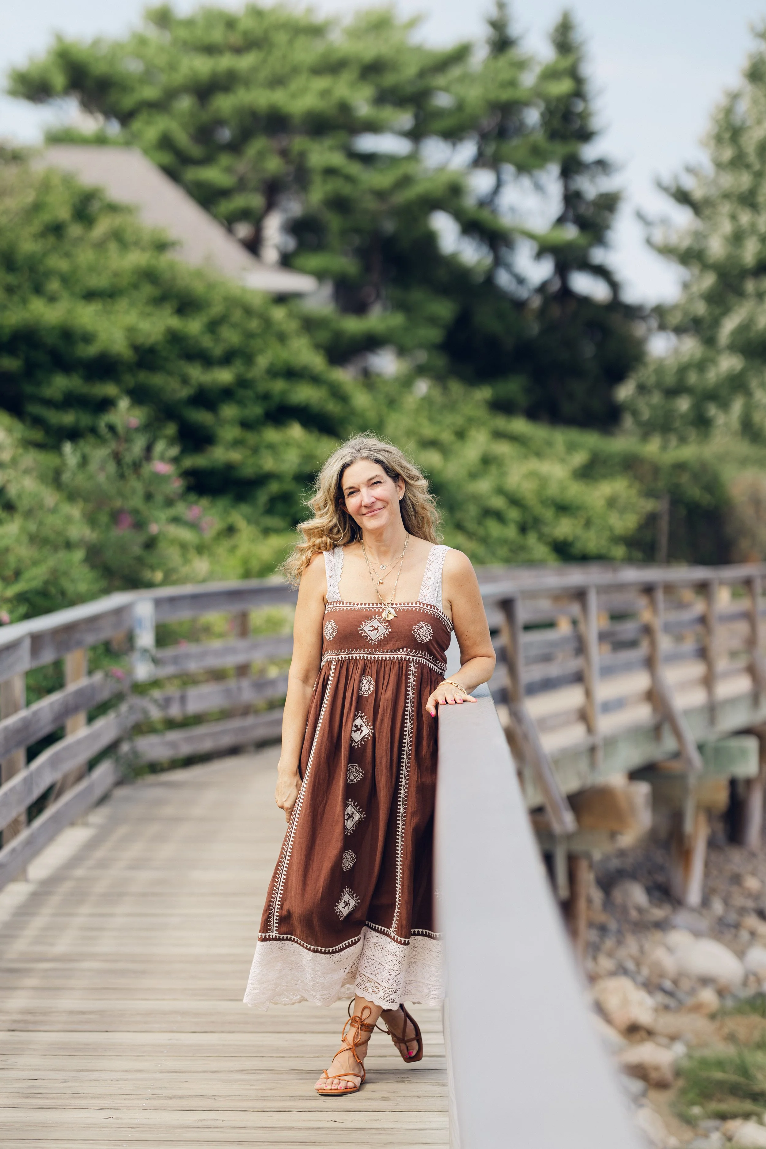 A woman with long, wavy blonde hair standing on a wooden bridge with a metal railing, smiling, wearing a brown boho-style dress and tan strappy sandals, surrounded by green trees and a gray house in the background.