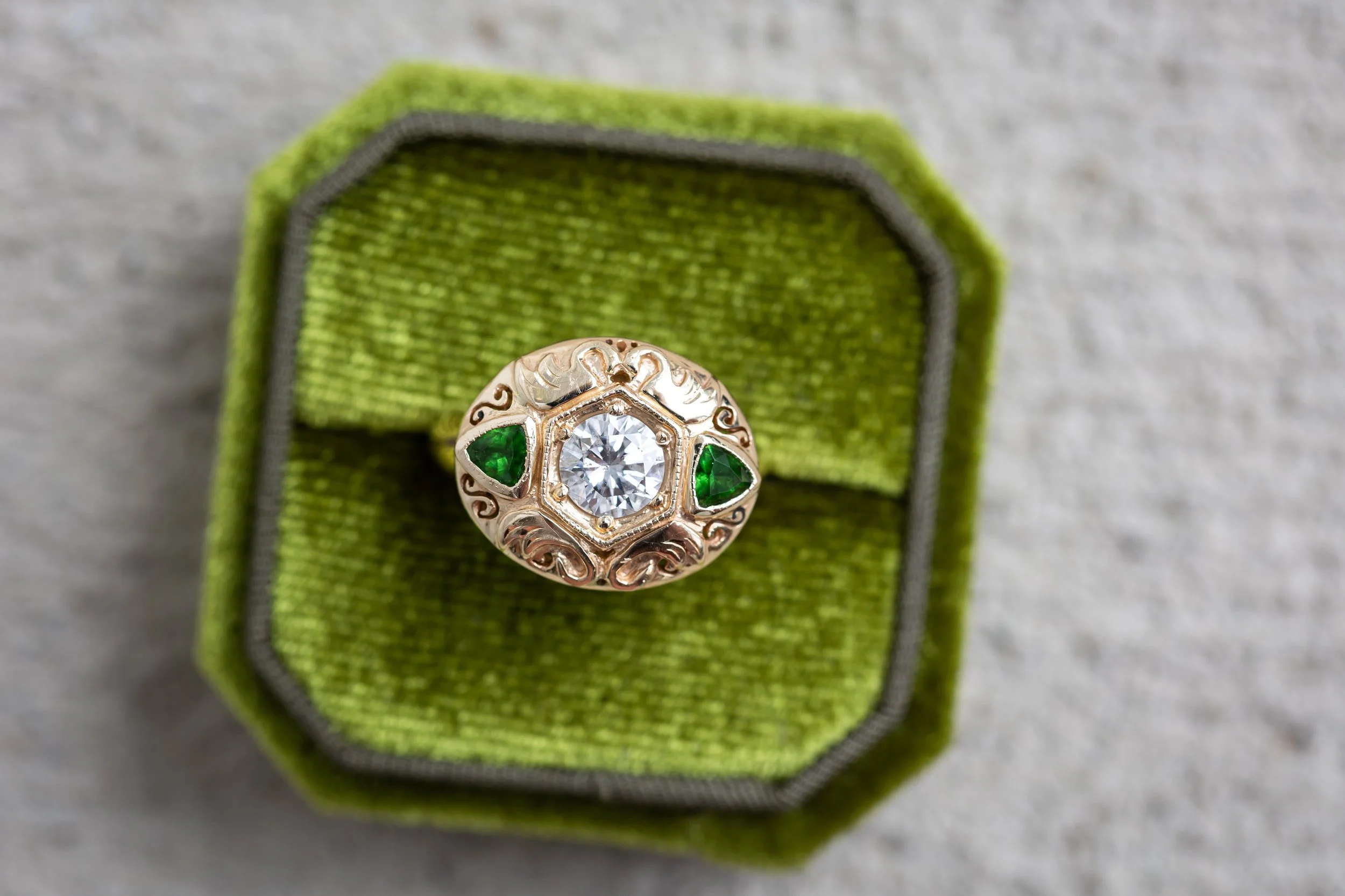 A decorative ring with a large central clear gemstone surrounded by ornate gold and green accents, placed on a green fabric-covered ring box.