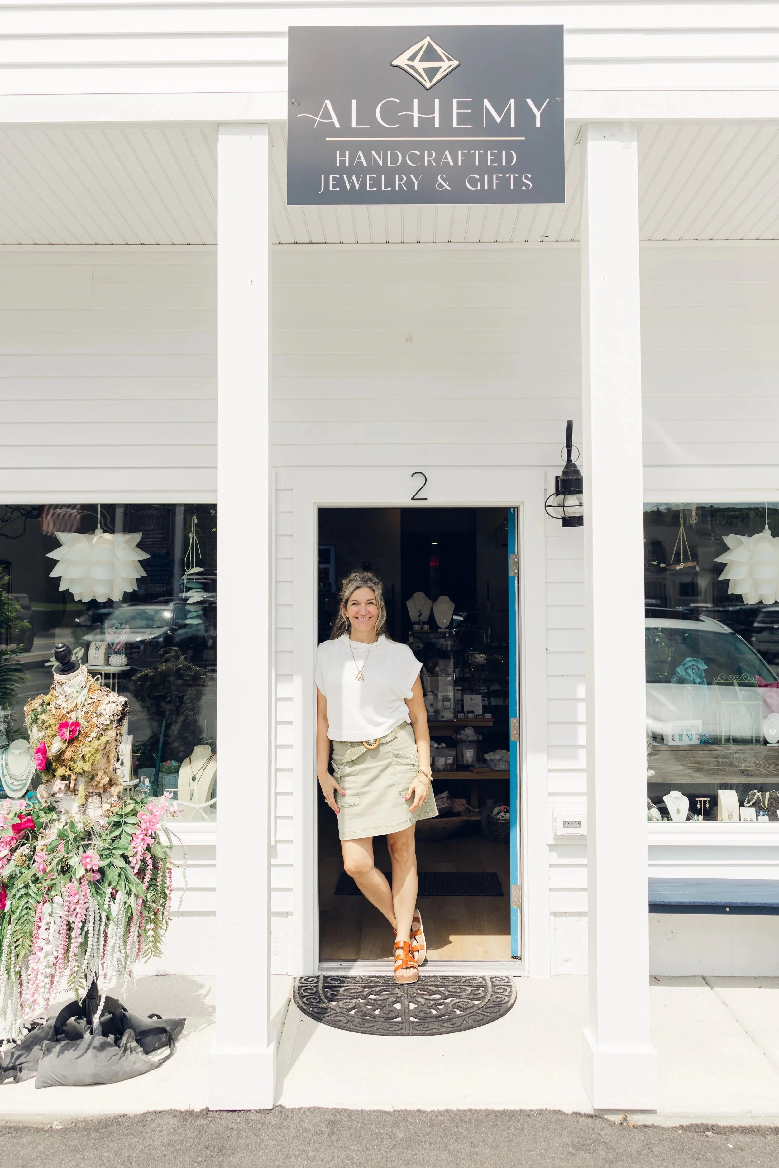 A woman standing in the doorway of a jewelry and gift shop named Alchemy, smiling, with a display of jewelry and gifts visible inside and outside.