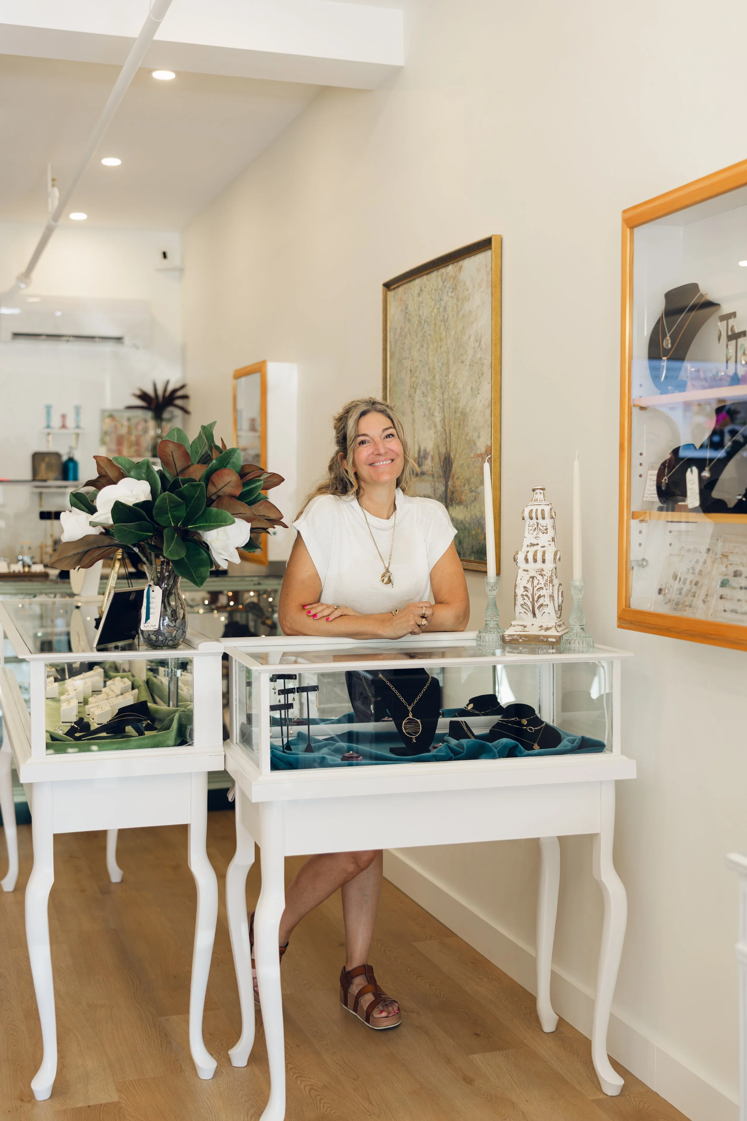 A woman standing behind a jewelry display counter in a boutique, smiling, with jewelry and decorative items on display around her, and framed artwork on the wall.
