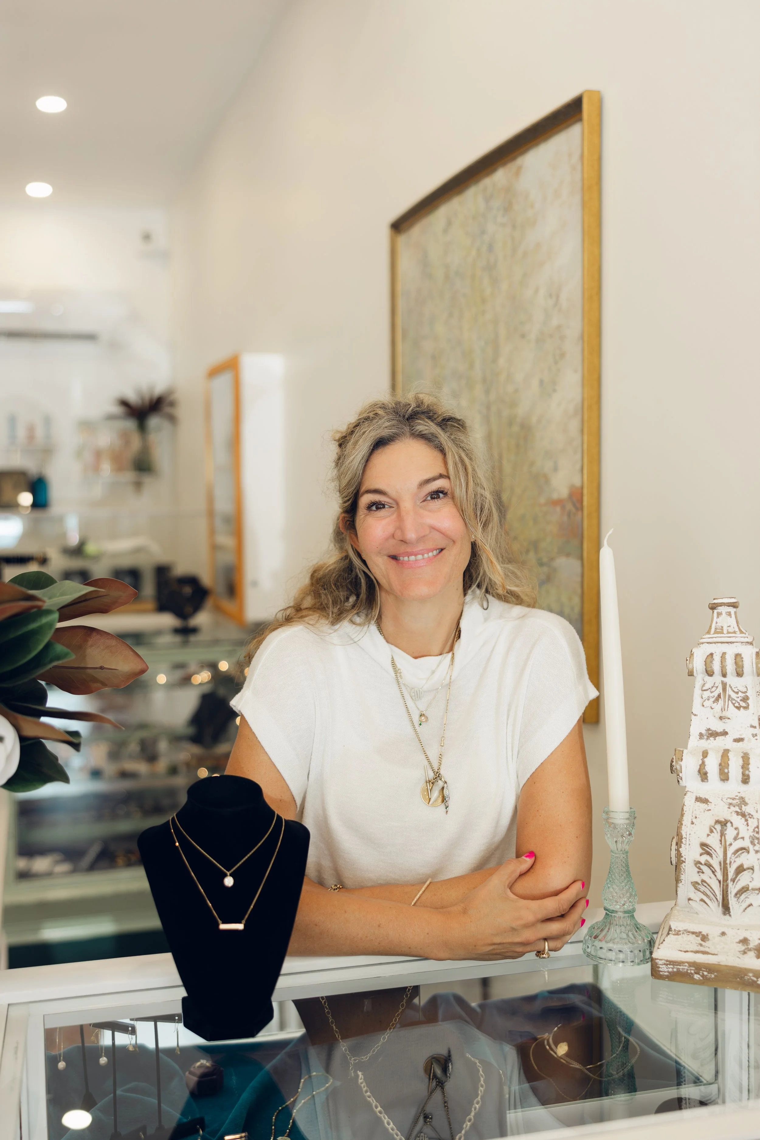 Smiling woman with blonde wavy hair, wearing a white top and layered gold necklaces, sits at a jewelry display counter in a store with jewelry on display, a candle, and a decorative urn on the table, and framed artwork behind her.