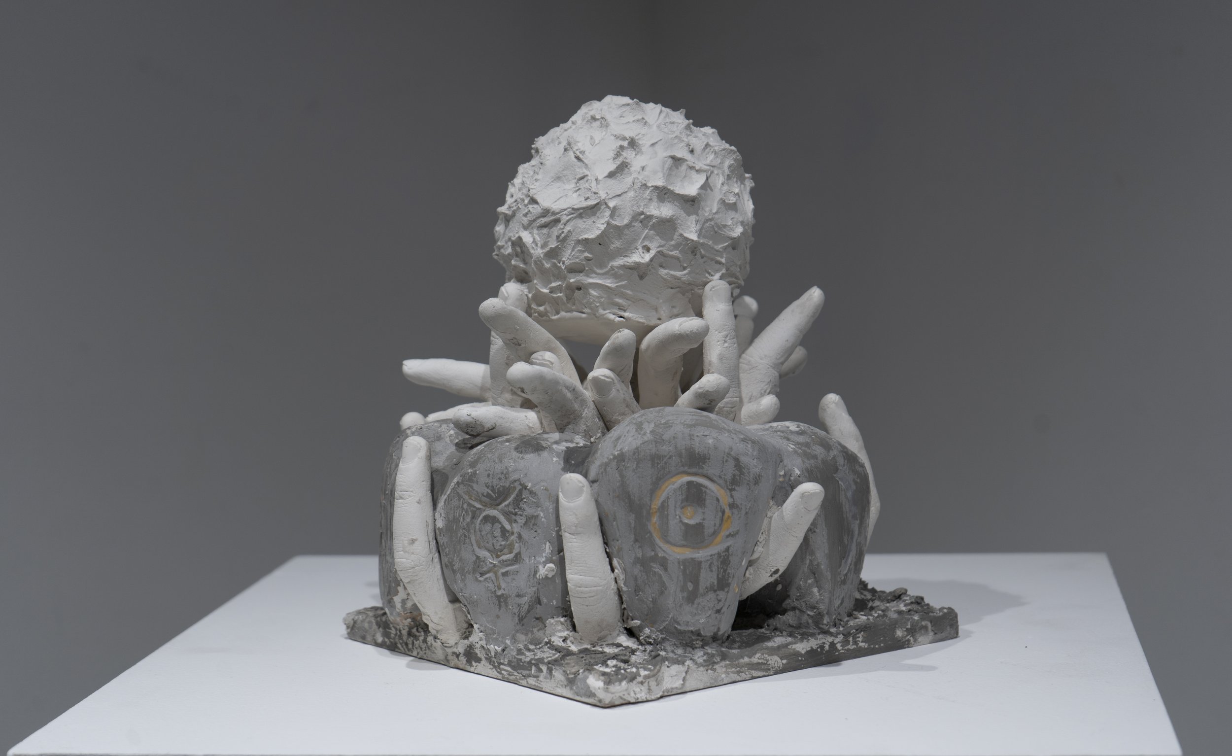 A sculpture of multiple white hands holding a textured, white, oval-shaped object against a gray background.