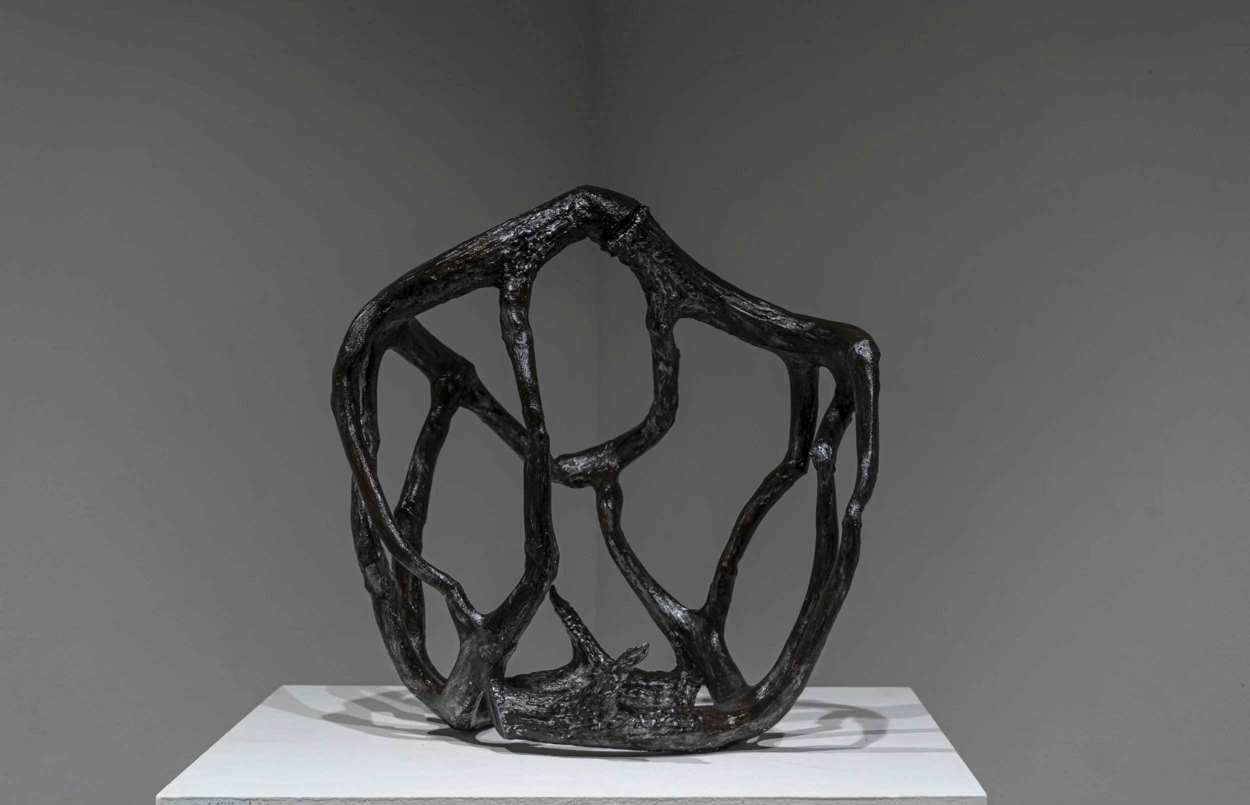 A modern sculpture made of dark, twisted metal resembling abstract interconnected figures, displayed on a white platform against a gray wall.