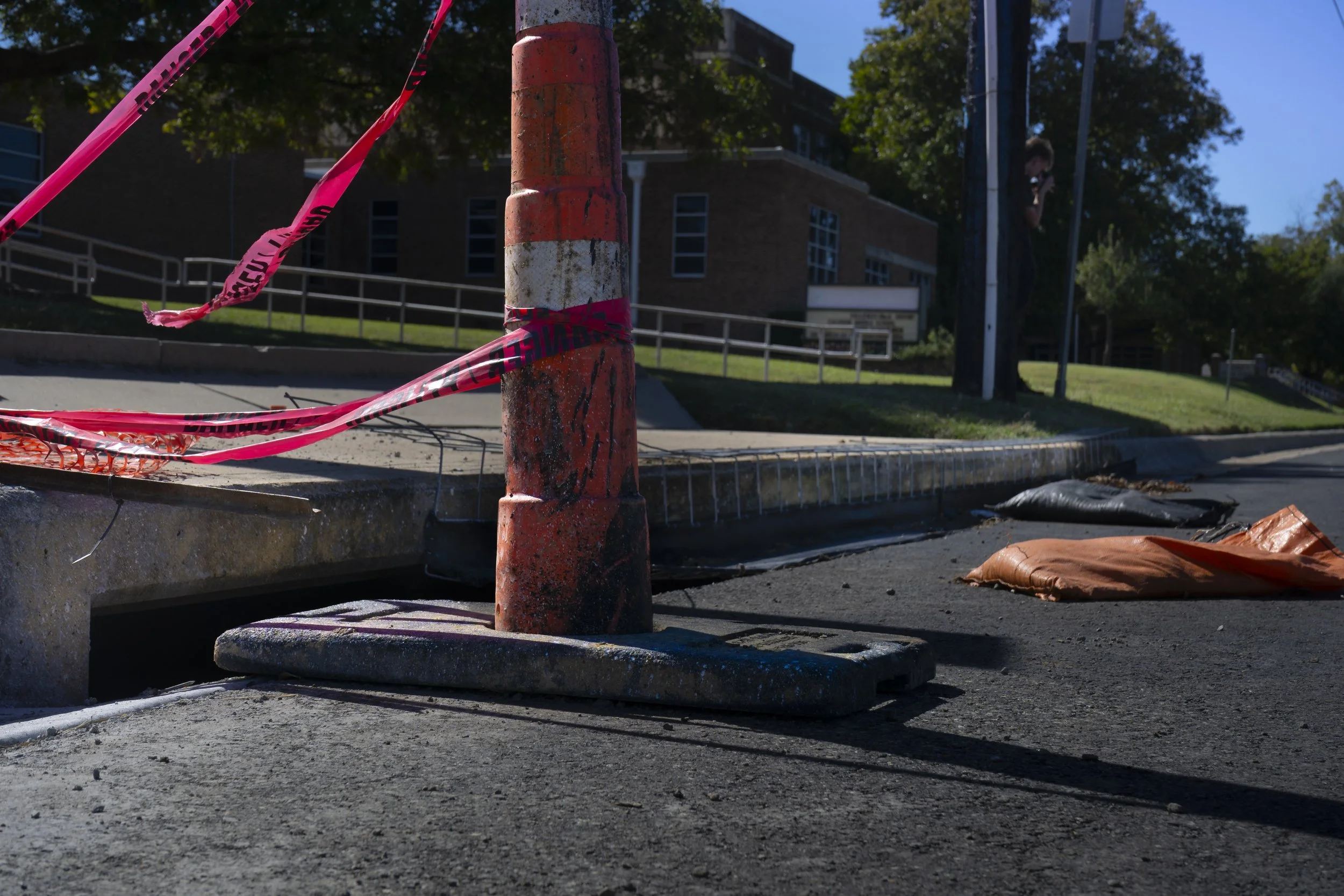 A construction site with a weathered orange safety cone wrapped with pink caution tape, surrounded by crushed orange safety barriers and materials on the ground. In the background are trees and a building, indicating an outdoor urban environment.