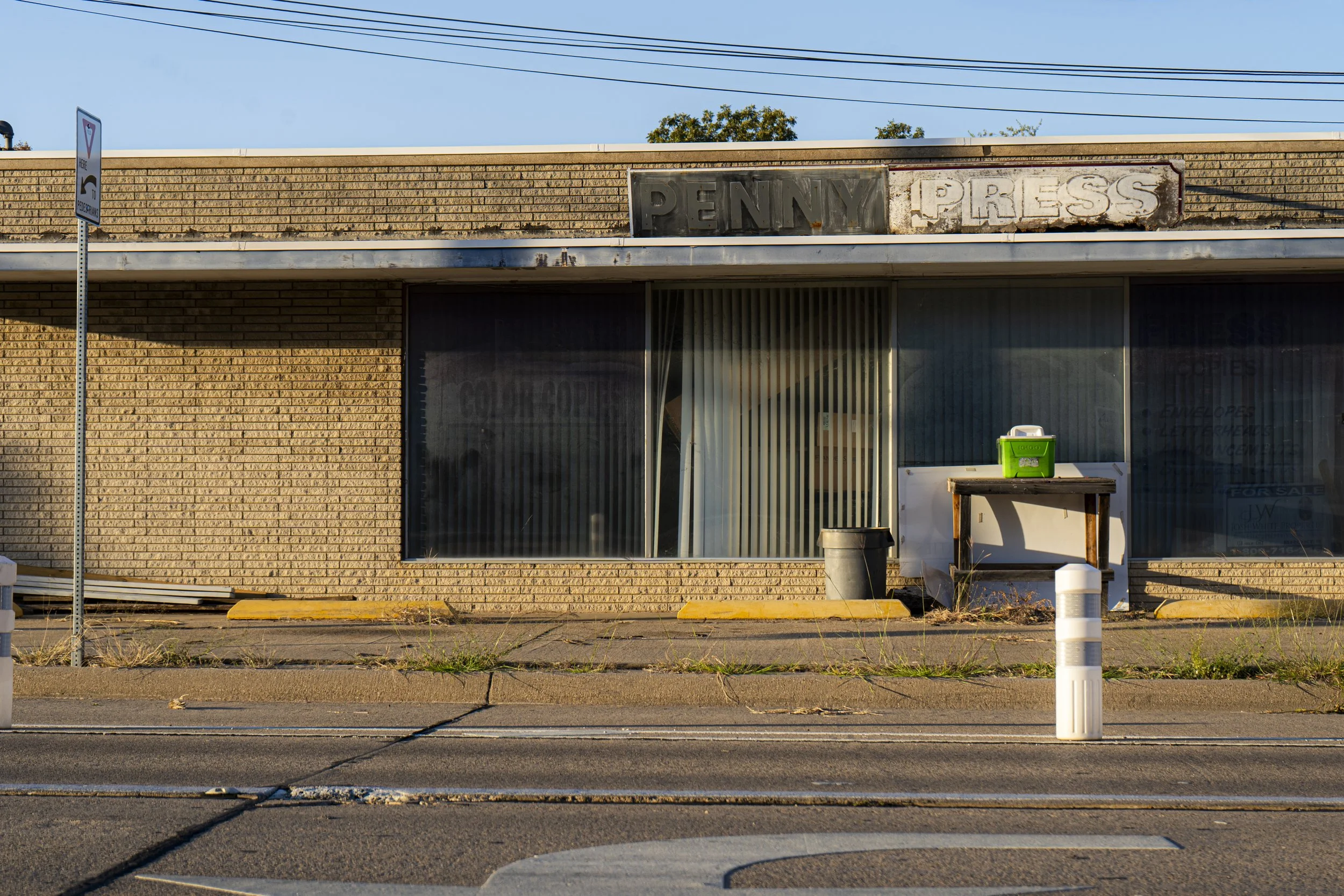 An empty storefront with a worn sign that reads 'PENNY PRESS' and a large window covered with vertical blinds. There are some miscellaneous objects outside, including a green box on a small table and a white cylindrical object, with a sidewalk and st