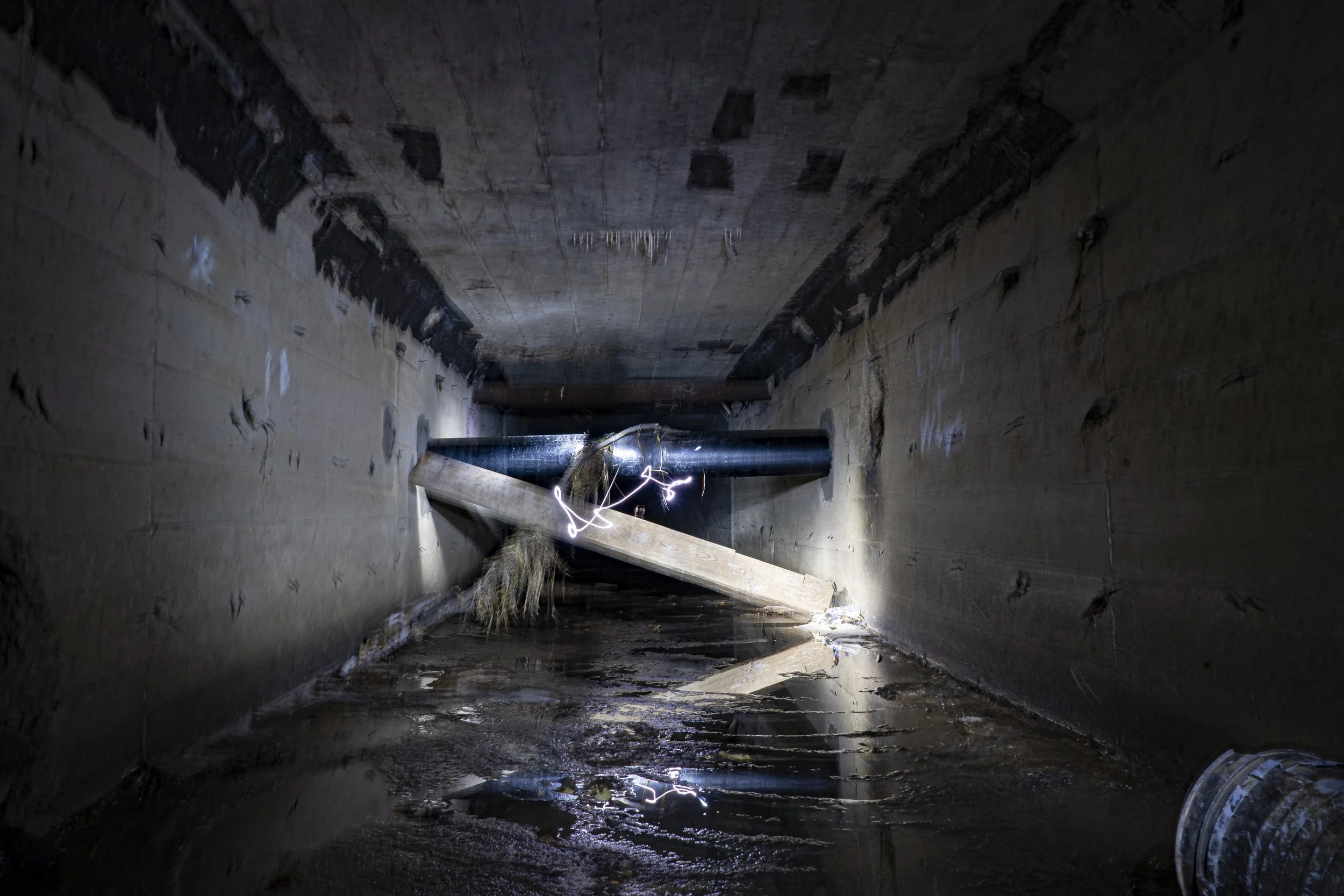 A dimly lit underground tunnel with water on the floor, a large black pipe resting across the tunnel, and construction or repair materials like a wooden board and electrical wires scattered inside.