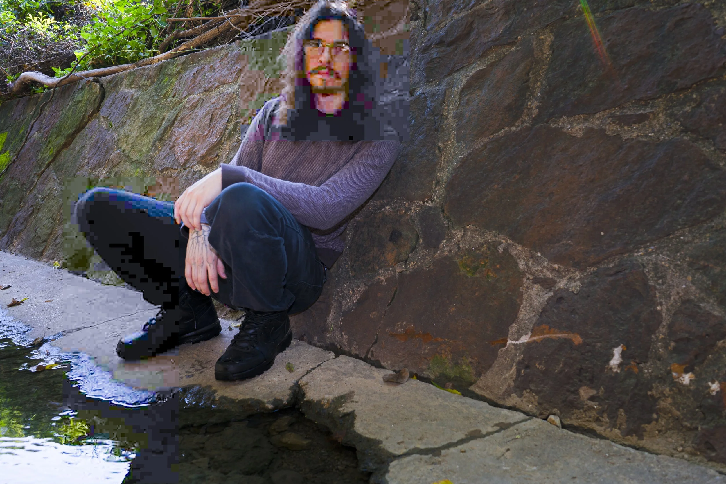 A person with glasses, dark hair, and a beard, sitting by a small body of water, wearing a purple sweater, dark pants, and black shoes, with a stone wall and green foliage in the background.
