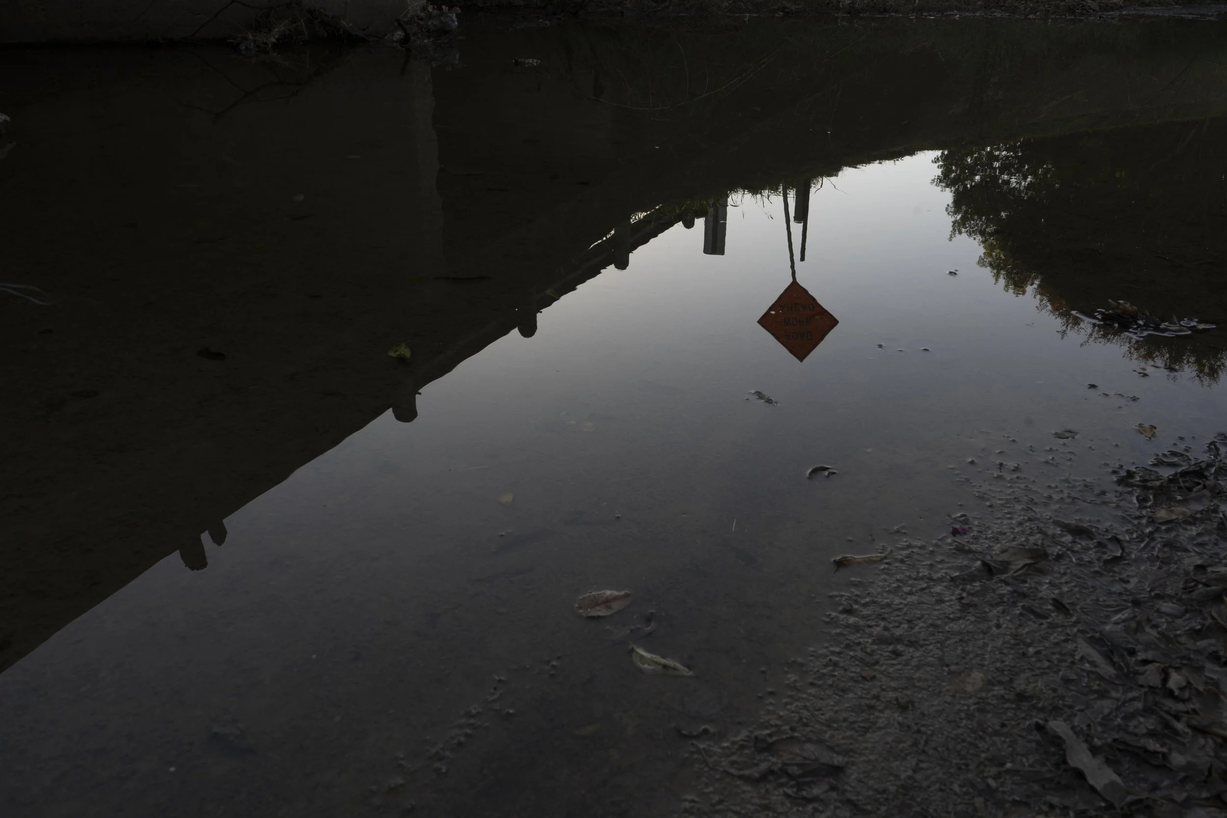 Reflection of a warning sign and a fence in a muddy puddle, with trees and sky reflected in the water.