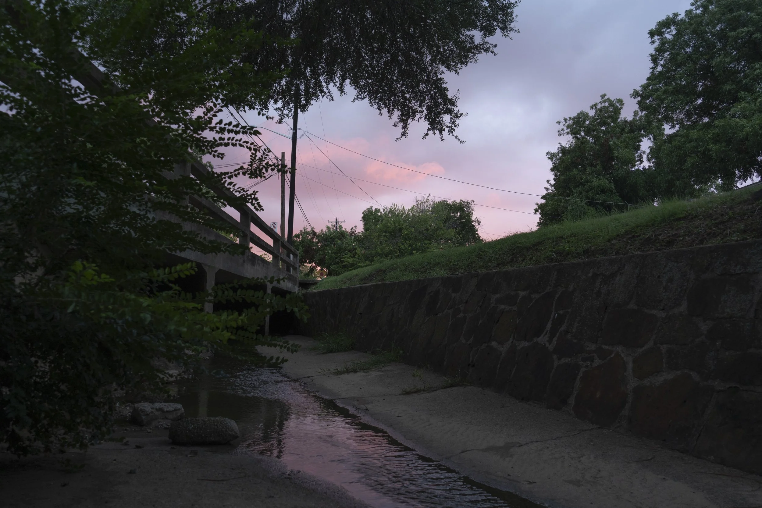 A small creek or stream flowing beneath a bridge in a wooded area during dusk or dawn. The sky has pink and purple hues, and trees with dense foliage surround the scene.