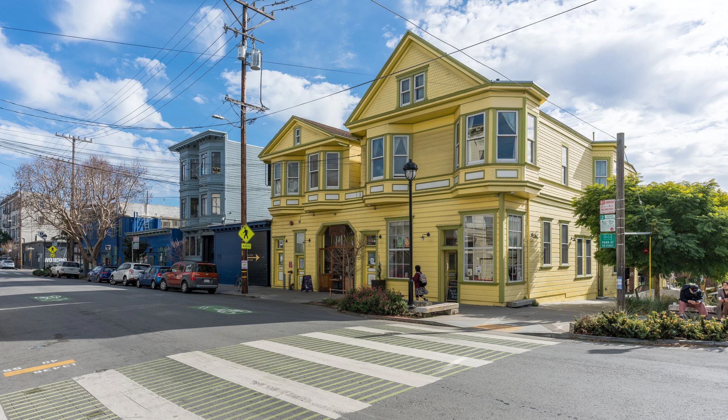 A yellow three-story building with bay windows on a city street, with a row of parked cars and a pedestrian crossing in front. Overhead power lines are visible against a blue sky with some clouds.
