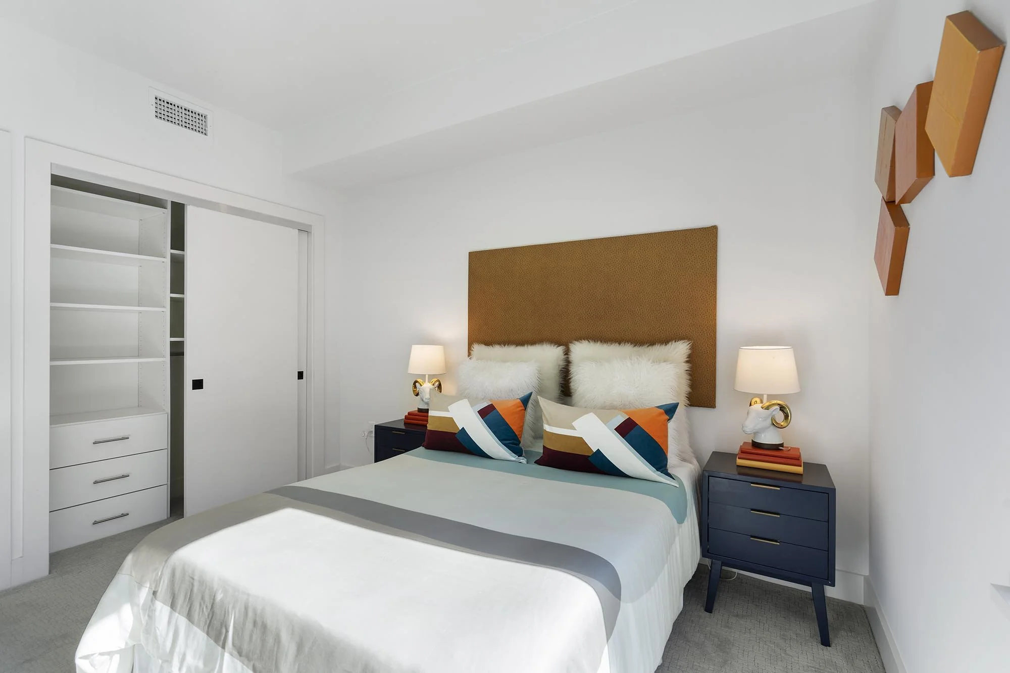 A modern bedroom with a white bed, colorful geometric pillows, a tan headboard, and two navy blue nightstands with white lamps featuring unique gold animal motifs. There is a partially open closet with white shelves and a sliding door, and a decorati