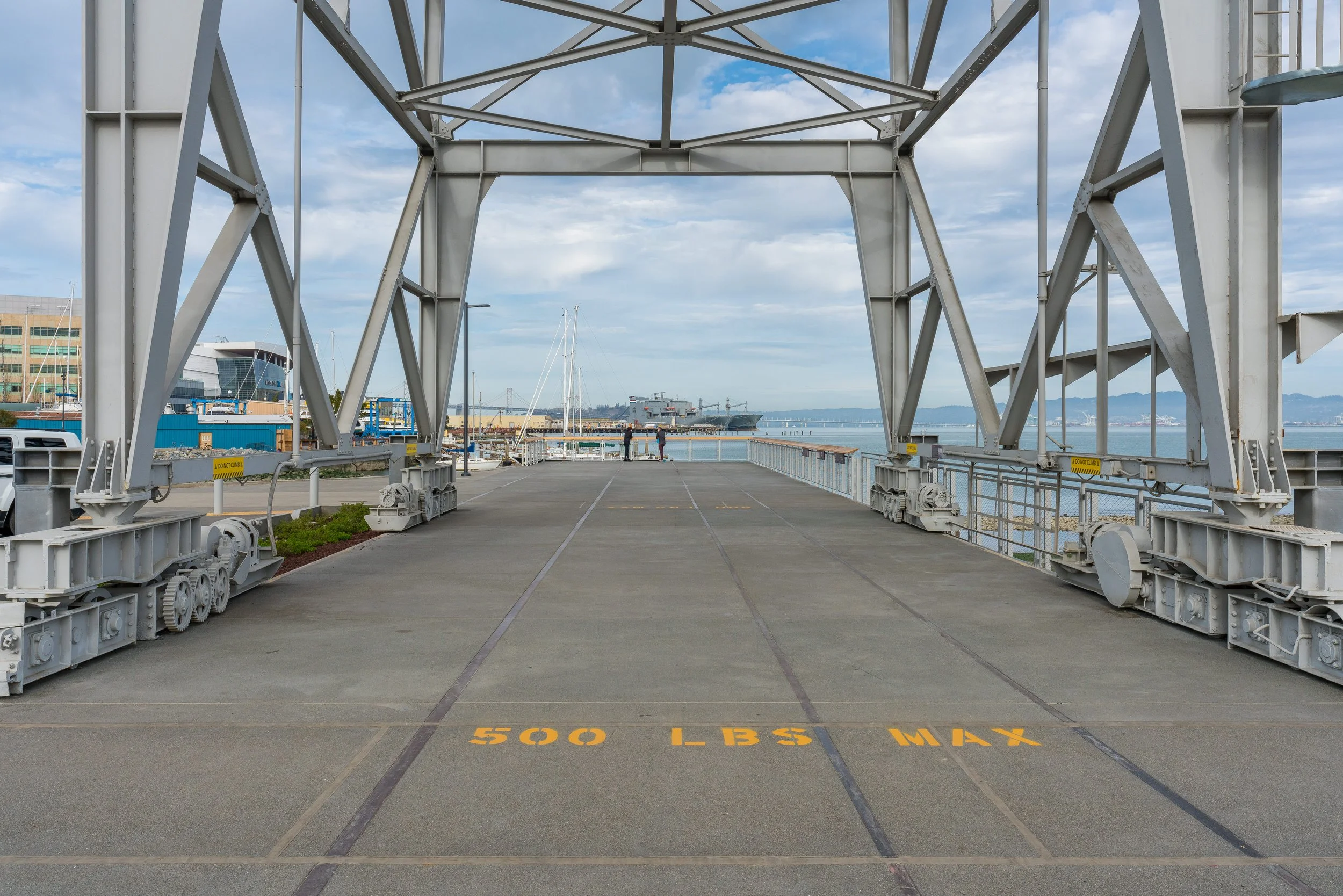 View from a ship lift at a harbor, showing large mechanical structures and a ship in the distance, with some buildings and a cloudy sky in the background.