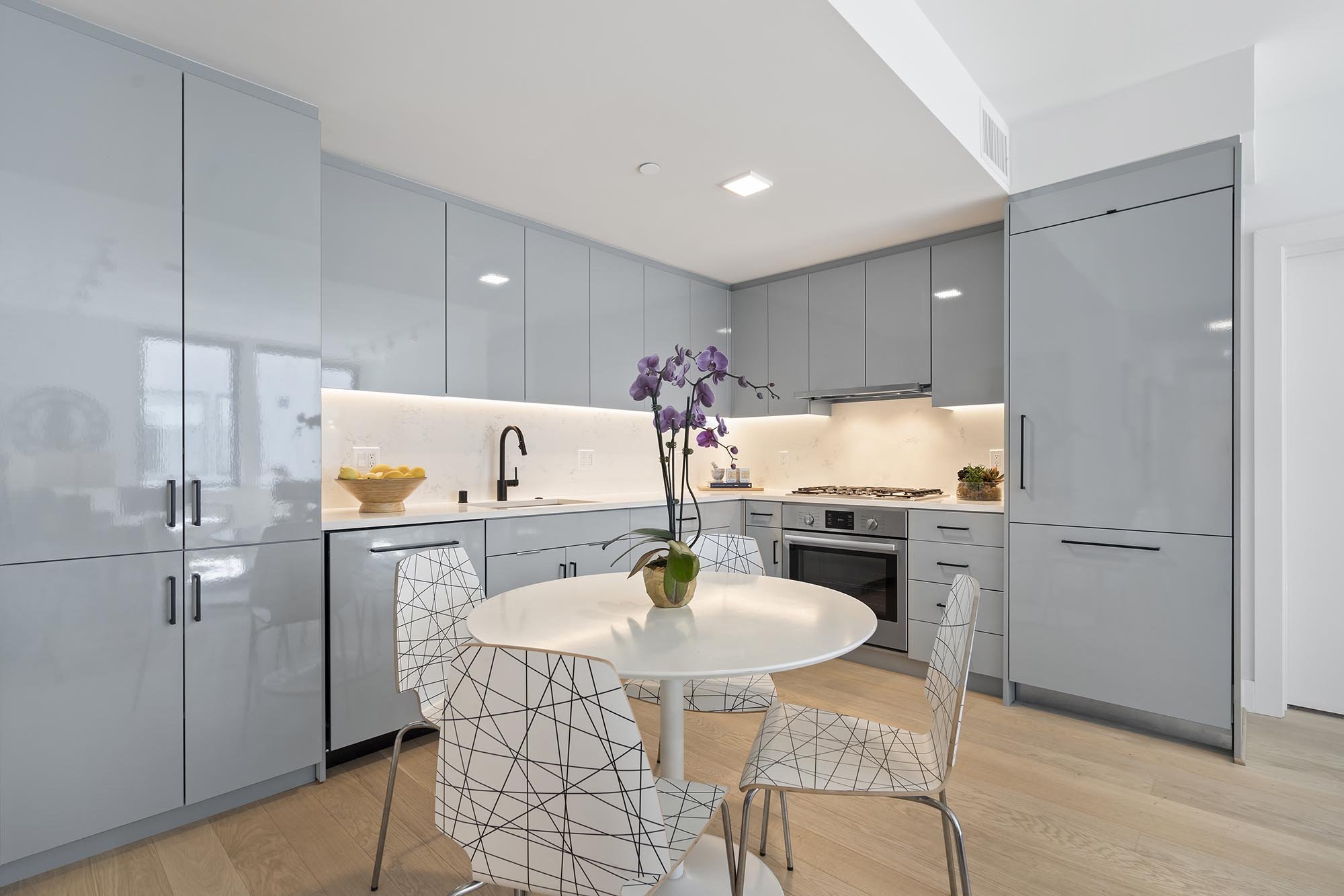 Modern kitchen with gray cabinetry, white countertops, a round white dining table with four wireframe chairs, a potted orchid, and built-in appliances in a bright space.