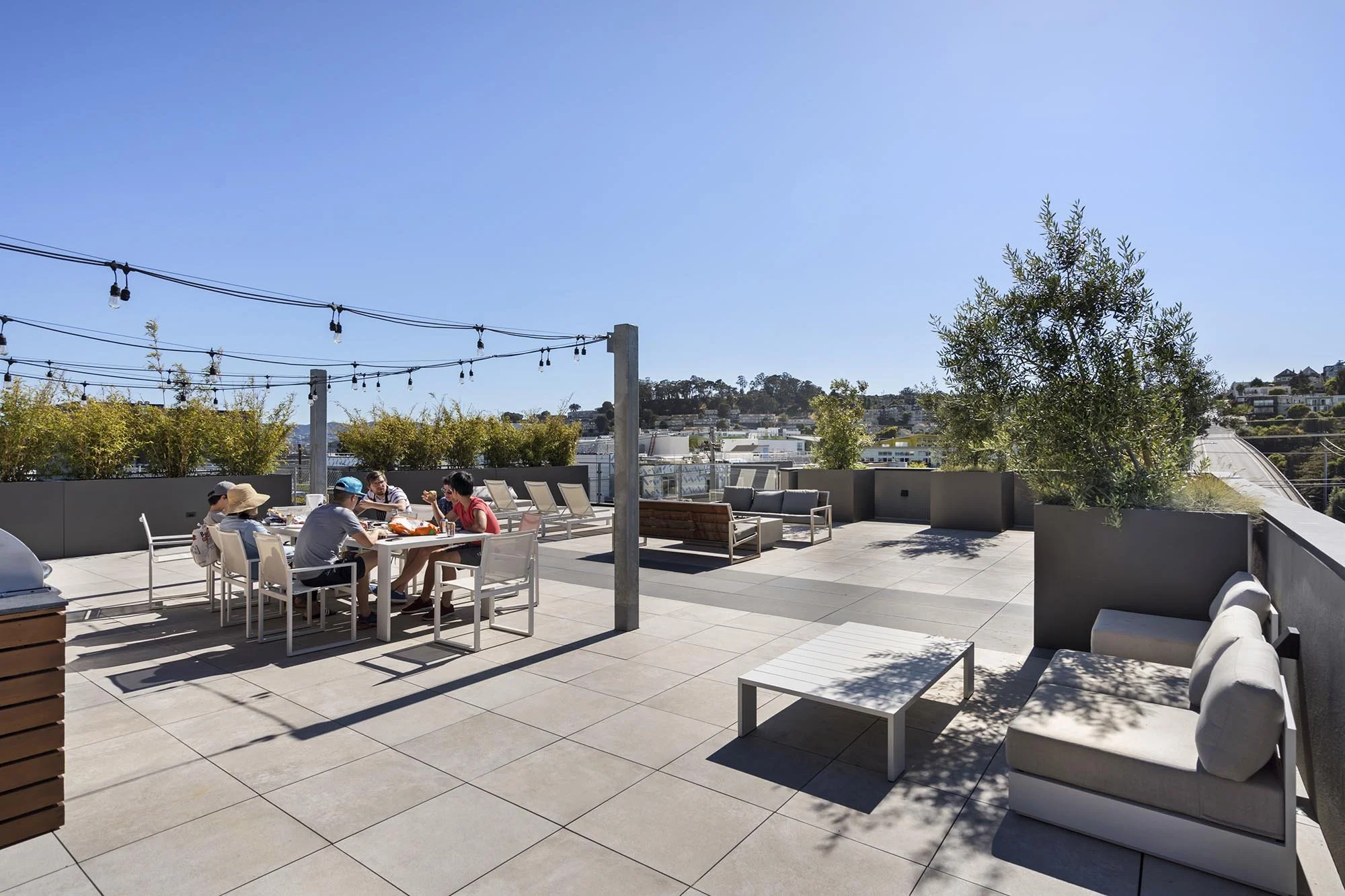 People dining at a rooftop patio with string lights, outdoor seating, potted trees, and a cityscape in the background under a clear blue sky.