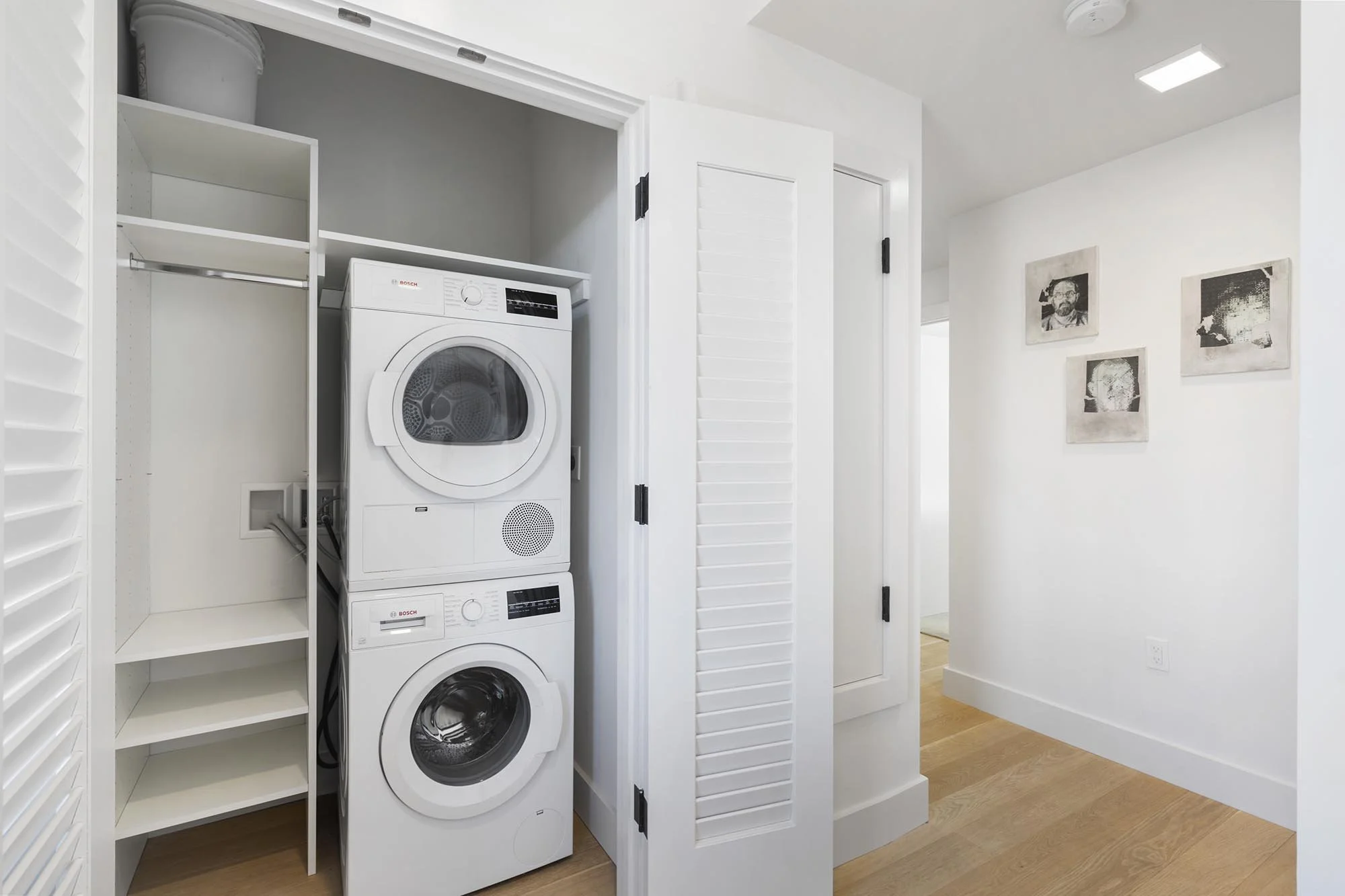 White laundry closet with stacked washing machine and dryer, open white shelves, and a storage door, with artwork on the adjacent wall.