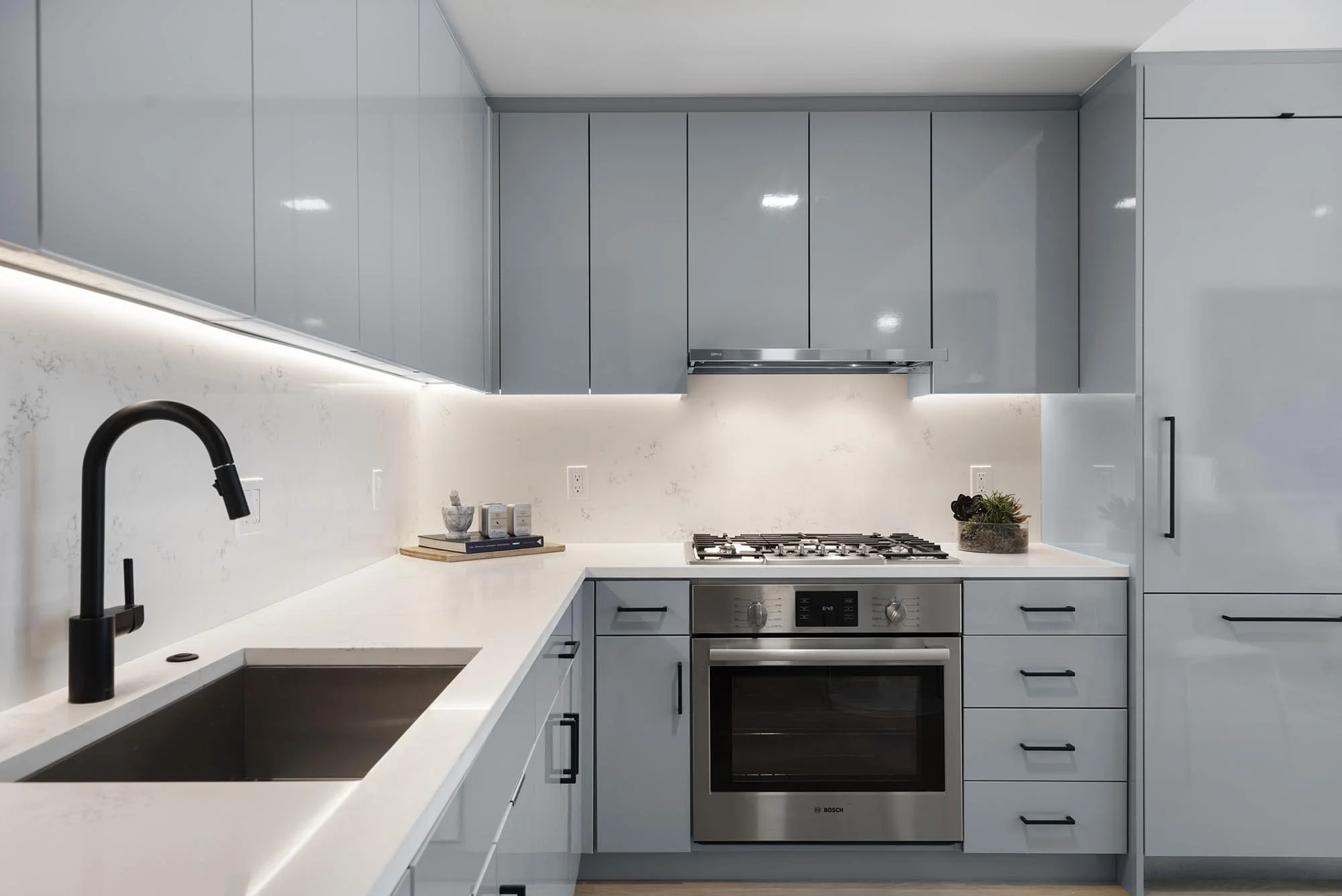 Modern kitchen with light grey cabinetry, white quartz countertop, and black fixtures. Contains a black kitchen faucet, a built-in oven, and a stovetop with an exhaust hood. Decorative items include a potted plant, a mortar and pestle, and a small st