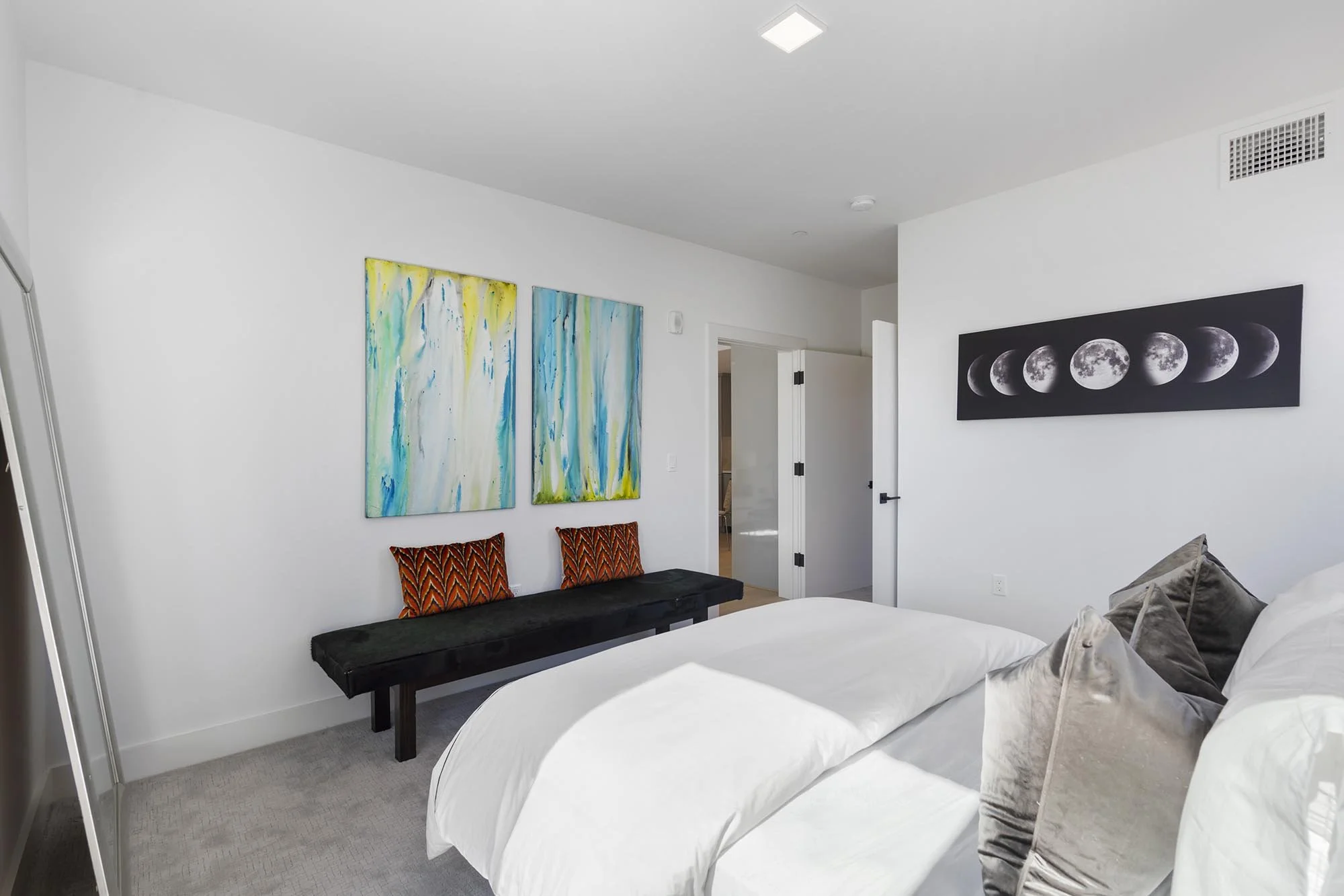 Modern bedroom with white walls, two artwork pieces, a black bench with two orange patterned pillows, a bed with multiple pillows, and a moon phase poster on the wall.