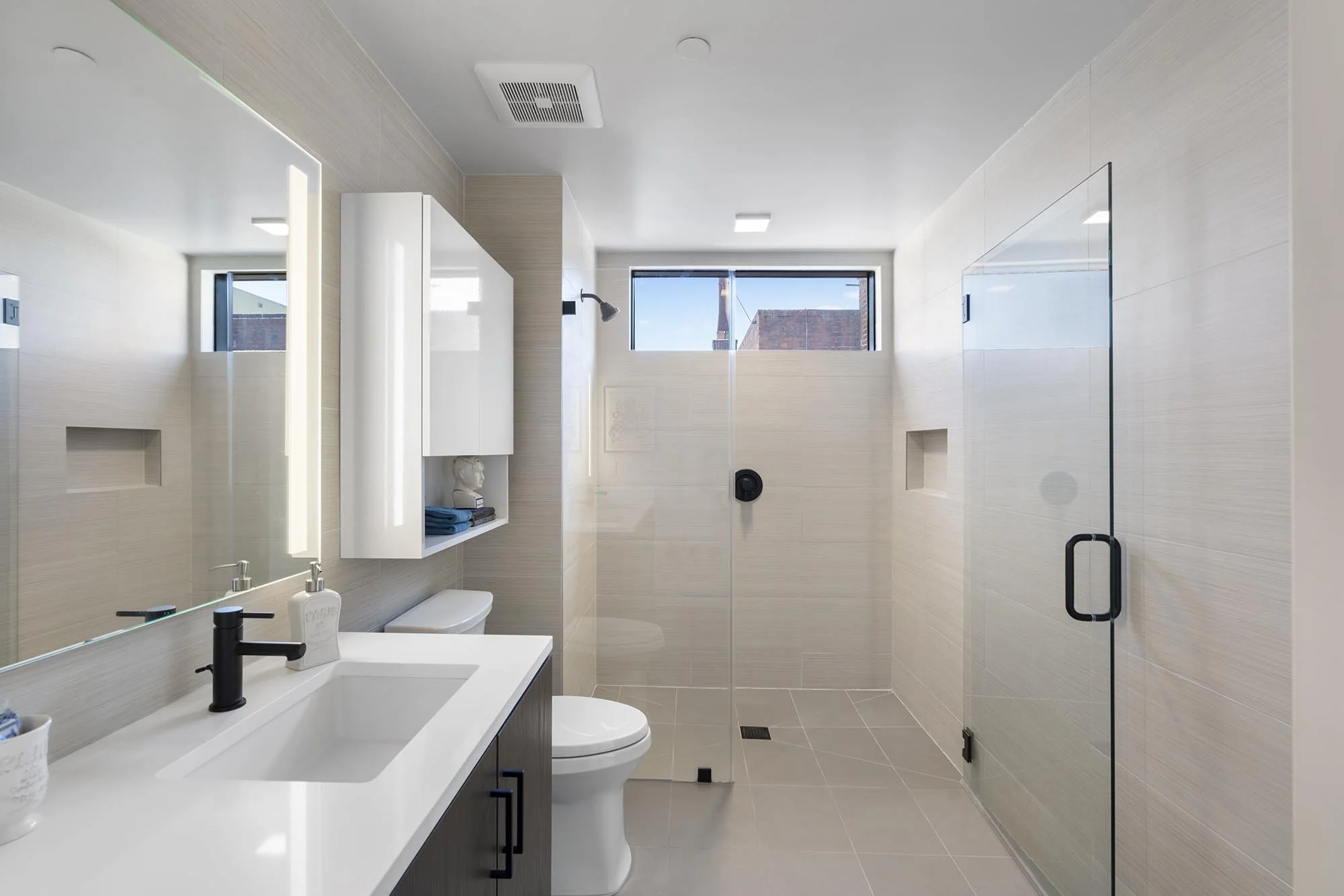 Modern bathroom with beige tiles, a white vanity with black fixtures, a wall-mounted white cabinet, a toilet, a glass shower with black hardware, two small windows, and natural light.