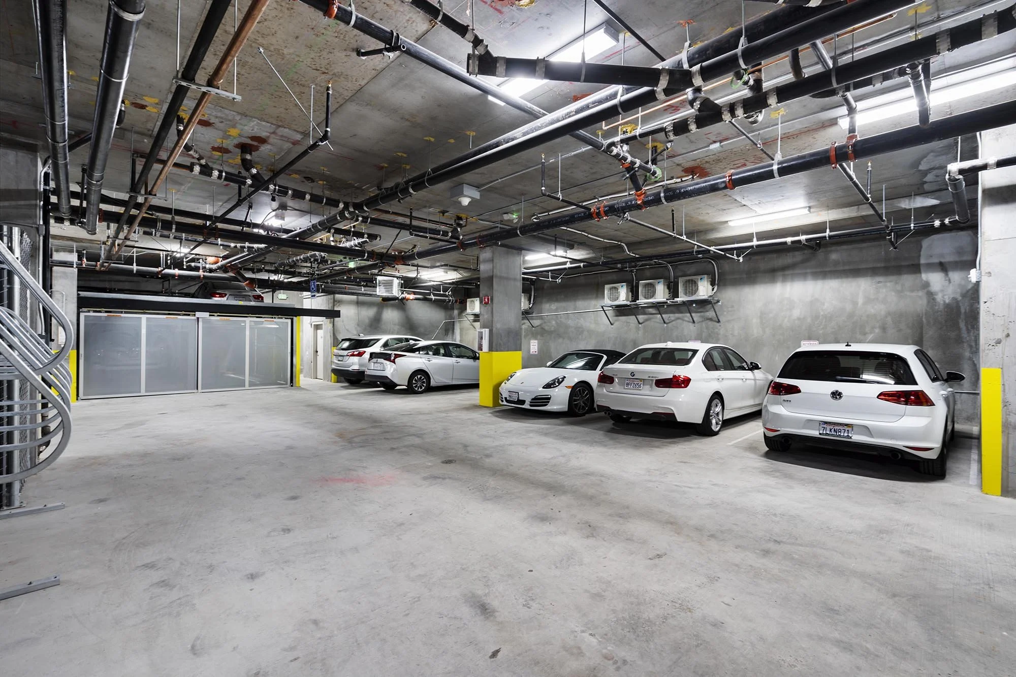 An underground parking garage with several cars parked, exposed concrete walls and ceiling, and visible piping and ductwork overhead.