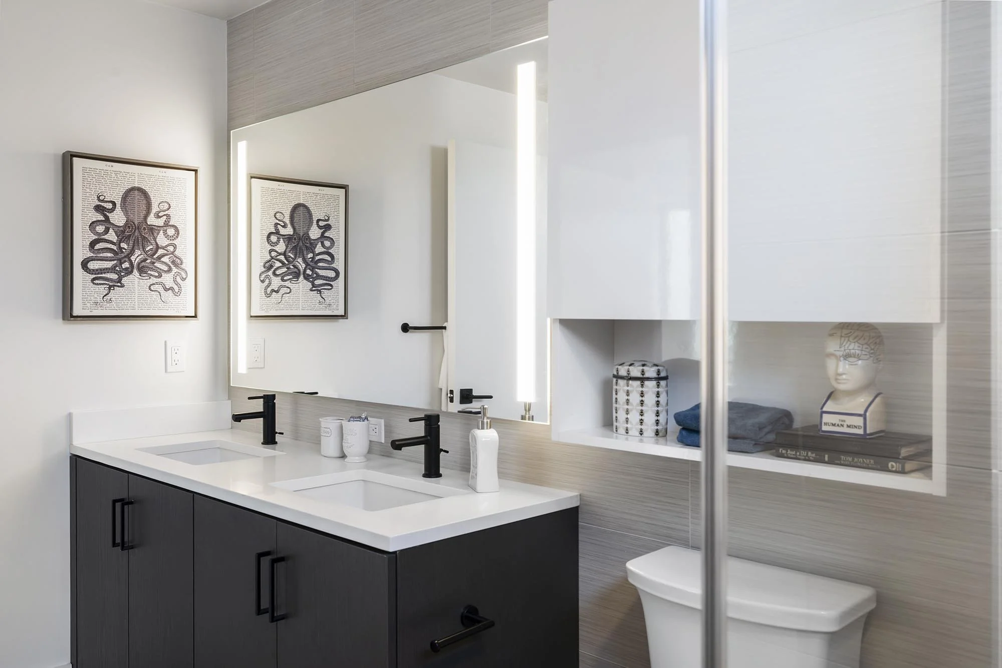 Modern bathroom with double vanity, black faucets, framed artwork of an octopus, open shelf with decorative items, and a mirror with lighting.