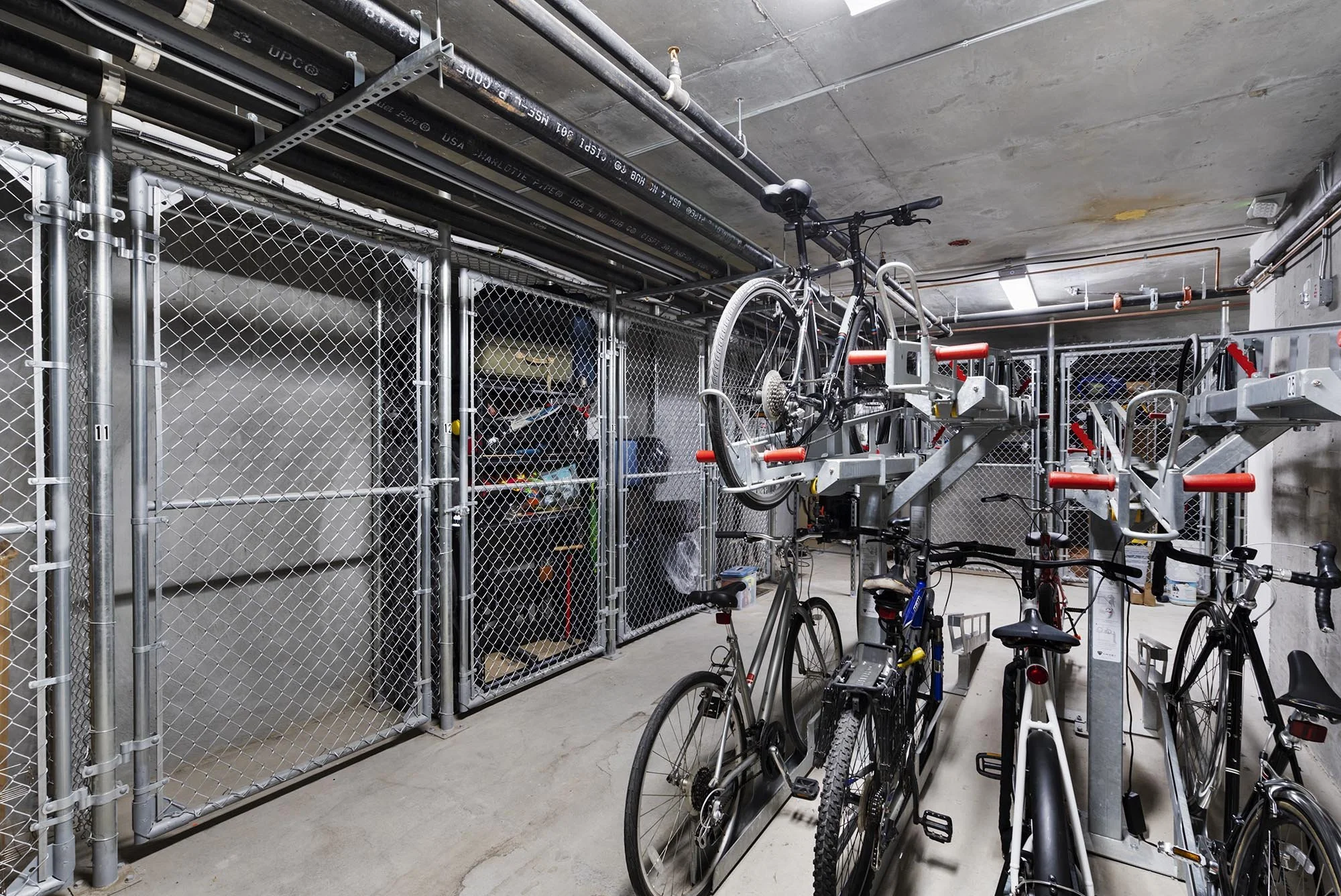 Indoor bike storage area with multiple bicycles parked on racks. The space is enclosed with metal chain-link fencing and has pipes along the ceiling.
