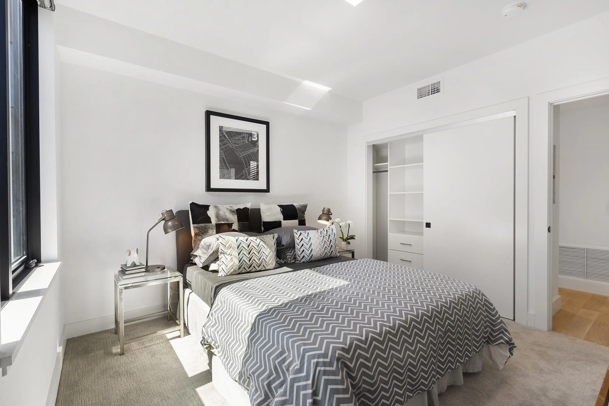 A modern bedroom with a large bed featuring a chevron patterned gray and white comforter and multiple pillows. There is a nightstand with a lamp, small decor, and a stack of books. The room has white walls, a framed black-and-white artwork, a window 