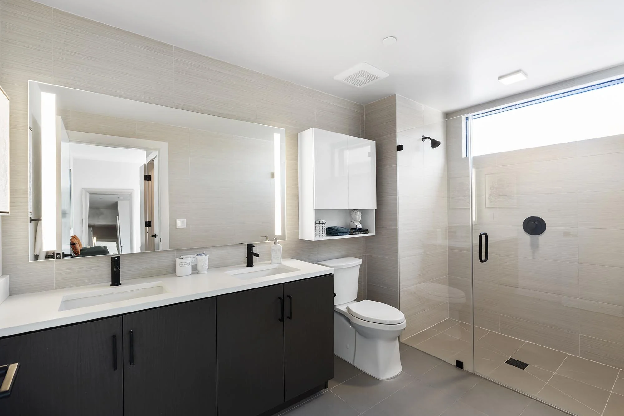 Modern bathroom with beige tiled walls, large mirror, white and black vanity, toilet, and a glass shower enclosure with a black handle, black showerhead, and small window near the ceiling.