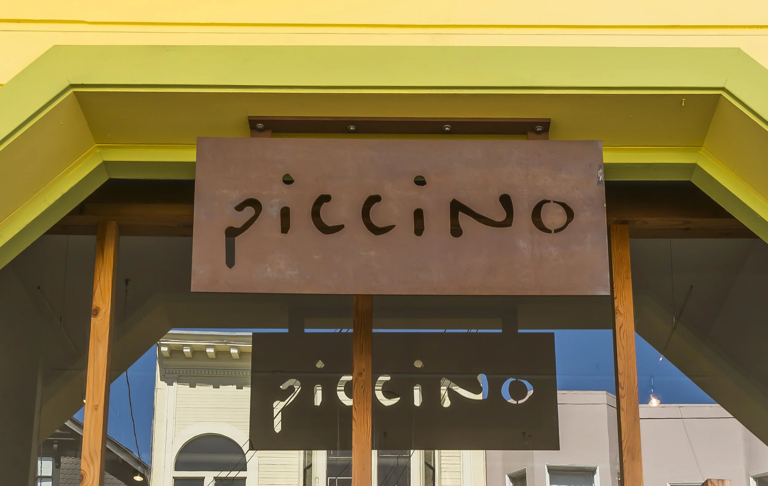 Storefront with a sign spelling 'Piccino' in cut-out letters hanging from a wooden frame over a walkway, with a building and sky in the background.