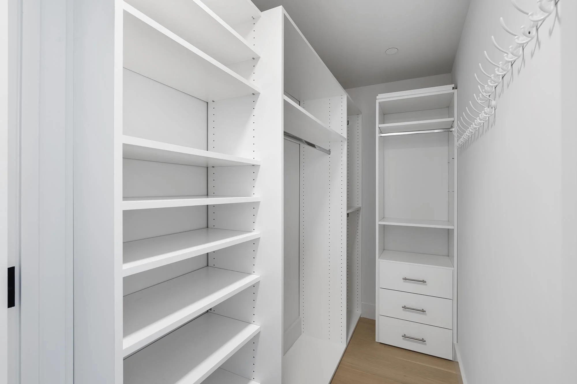 Empty white walk-in closet with built-in shelves, drawers, and hanging rods, and hardwood flooring.